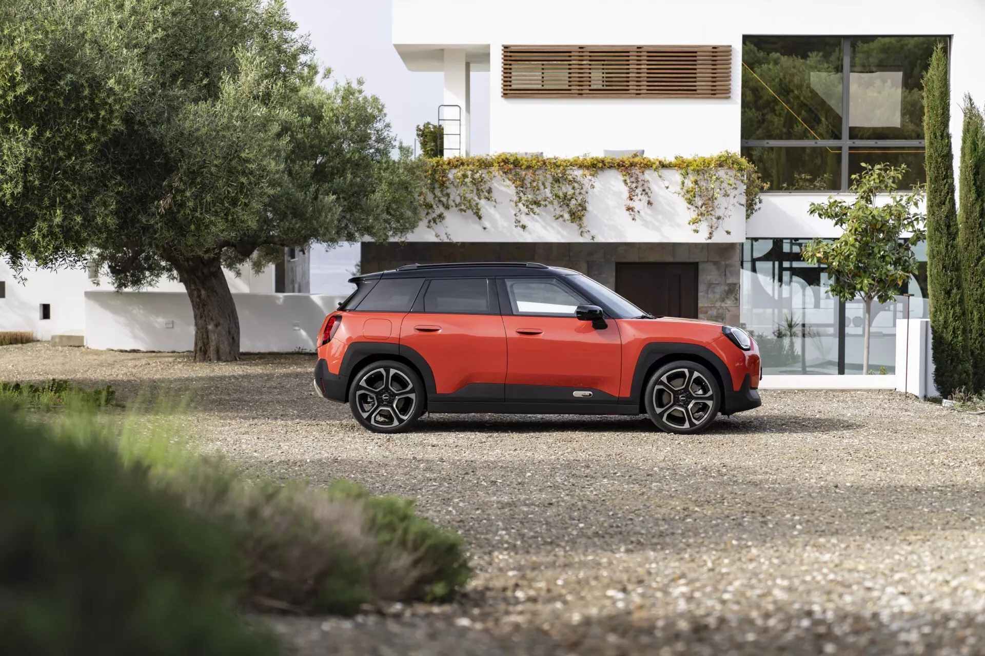 SUV Mini Aceman 2024 dans un audacieux schéma de couleurs orange-rouge et noir garé devant une maison contemporaine, montrant ses profils avant et latéral avec des lignes de design distinctives, des phares LED, des roues à plusieurs branches et une haute garde au sol.