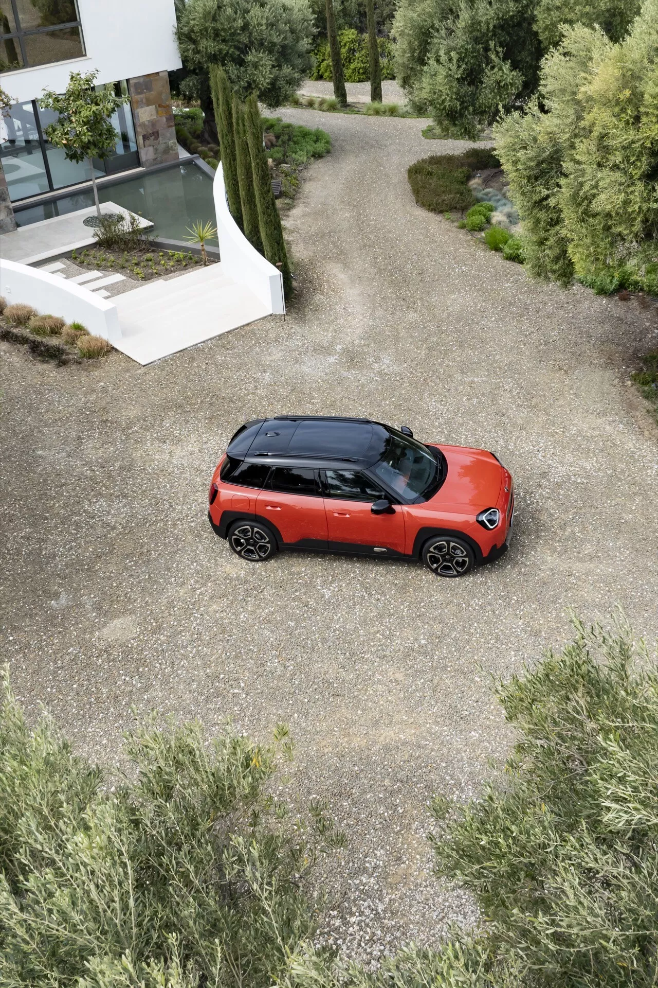 Vue aérienne du Mini Aceman (J05) 2024 en rouge vif avec toit noir garé sur un chemin de gravier menant à un bâtiment moderne parmi la verdure, présentant un design de SUV compact, peinture bicolore, toit panoramique et jantes en alliage à multiples branches.