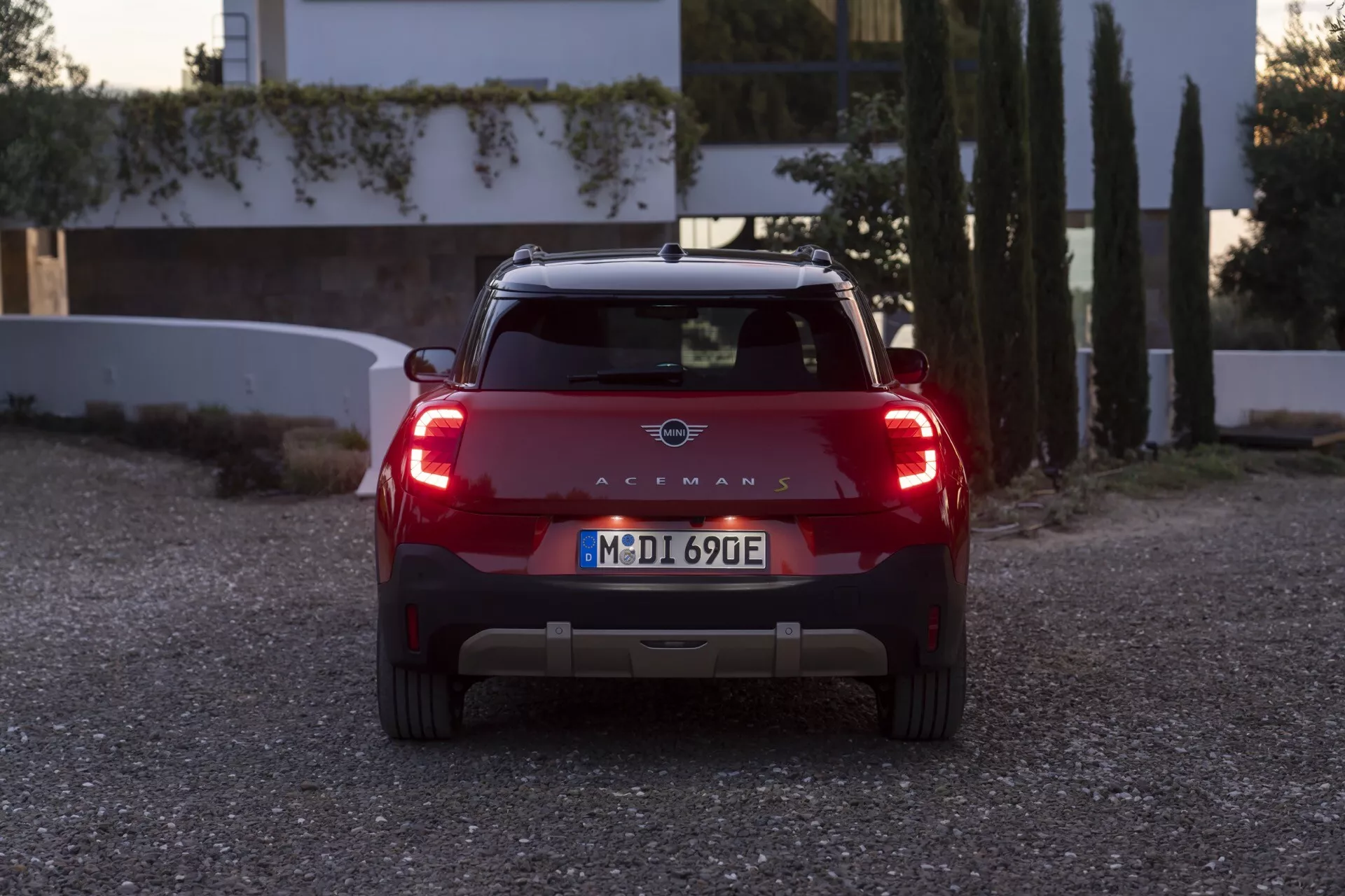 Vue arrière du Mini Aceman 2024 (J05) pendant un lever ou coucher de soleil pittoresque, mettant en avant les feux arrière illuminés, la couleur rouge vibrante et les caractéristiques technologiques durables contre un fond de nature et d'architecture