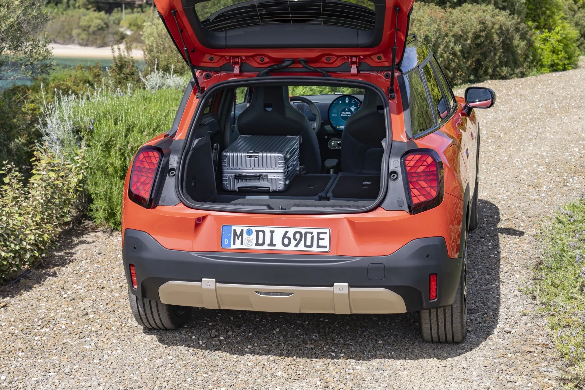 Vue arrière d'une hatchback orange Mini Aceman (J05) de 2024, se distinguant par son groupe motopropulseur électrique ou hybride, avec le coffre ouvert affichant un grand composant du groupe motopropulseur, possiblement à des fins éducatives ou de service, stationnée sur une surface de gravier entourée de verdure.