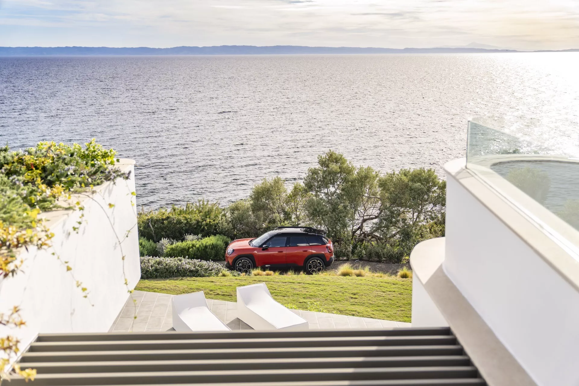 Mini Aceman 2024 (J05) garée sur l'herbe près de la côte avec un arrière-plan structurel moderne et un escalier descendant, mettant en avant un design élégant rouge, des feux arrière à LED et des jantes en alliage