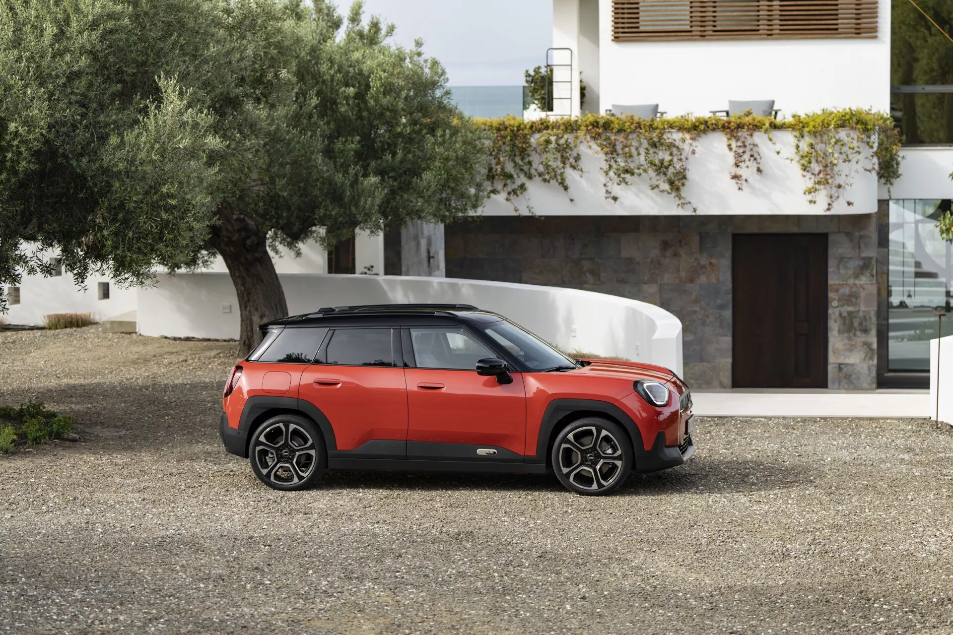 Mini Aceman 2024 (J05) en rouge avec toit noir, présentant son design de SUV compact sous un angle avant de trois quarts sur une surface de gravier, mettant en avant la grille frontale, les phares élégants, les roues sportives et le profil latéral distinctif sur fond de résidence contemporaine