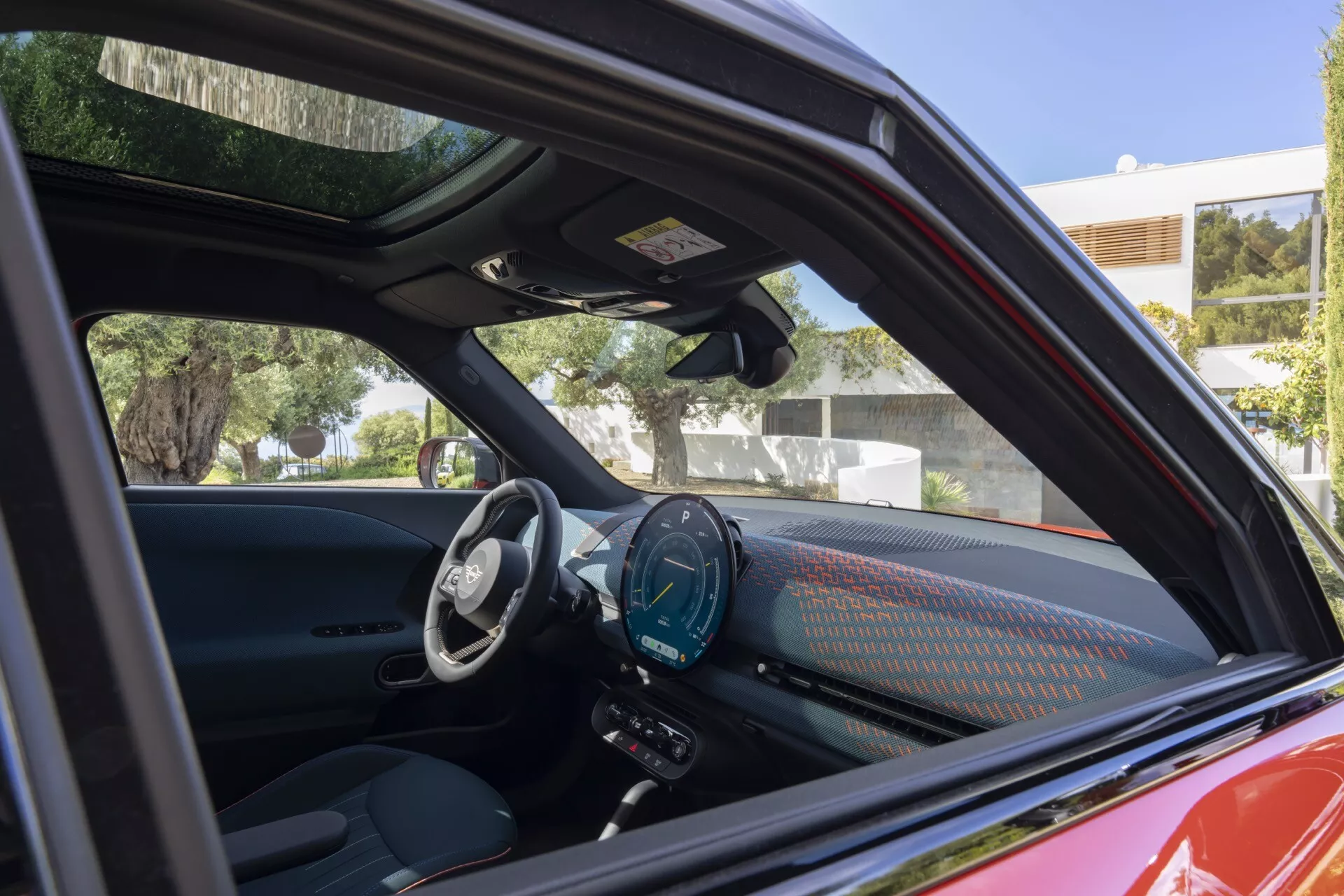 Vue intérieure du Mini Aceman 2024 (J05) à travers la fenêtre ouverte côté conducteur, affichant un design moderne avec un volant rempli de technologies, un tableau de bord numérique et un grand écran tactile d’infodivertissement avec navigation, sièges en cuir premium et un panneau de porte élégant avec contrôles dans un cadre de cabine luxueux.