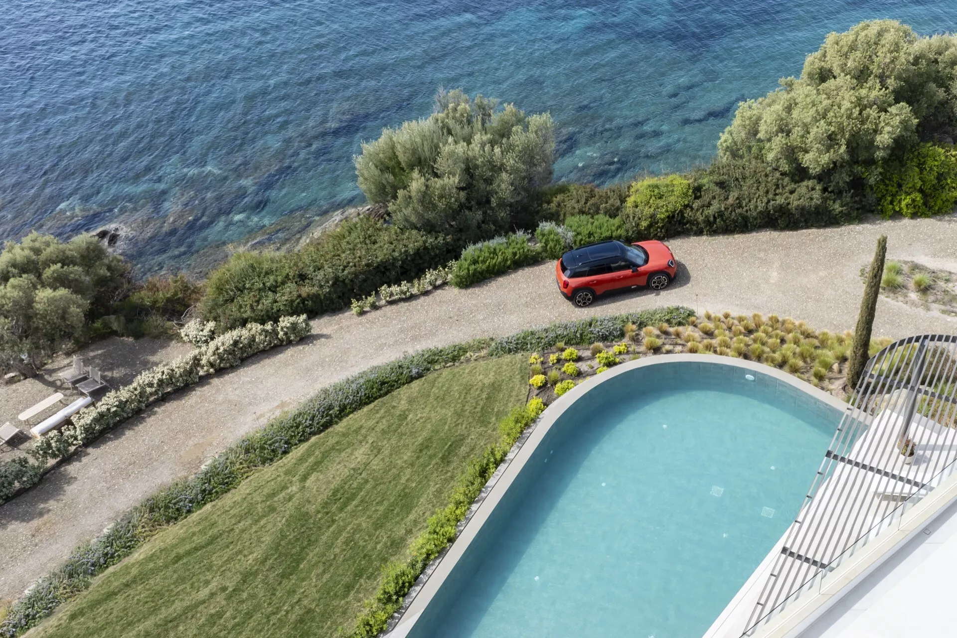Mini Aceman (J05) de 2024 en rouge, garée sur une propriété de luxe avec vue sur une piscine et un plan d'eau, mettant en avant son design aérodynamique et sa finition brillante