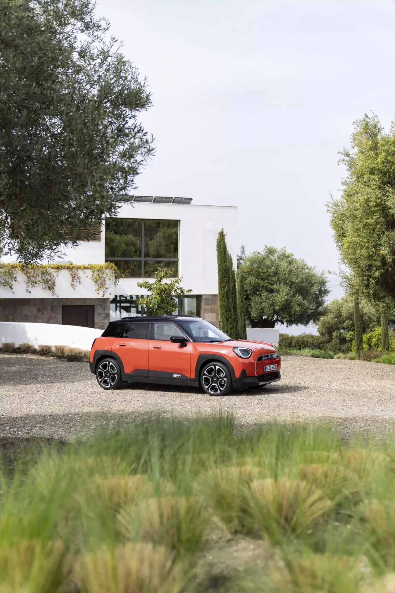 Mini Aceman 2024 (J05) SUV compact quatre portes sur gravier, montrant vue de face et côté avec maison contemporaine et verdure en arrière-plan, avec toit contrasté, phares LED modernes et éventuelle personnalisation haut de gamme