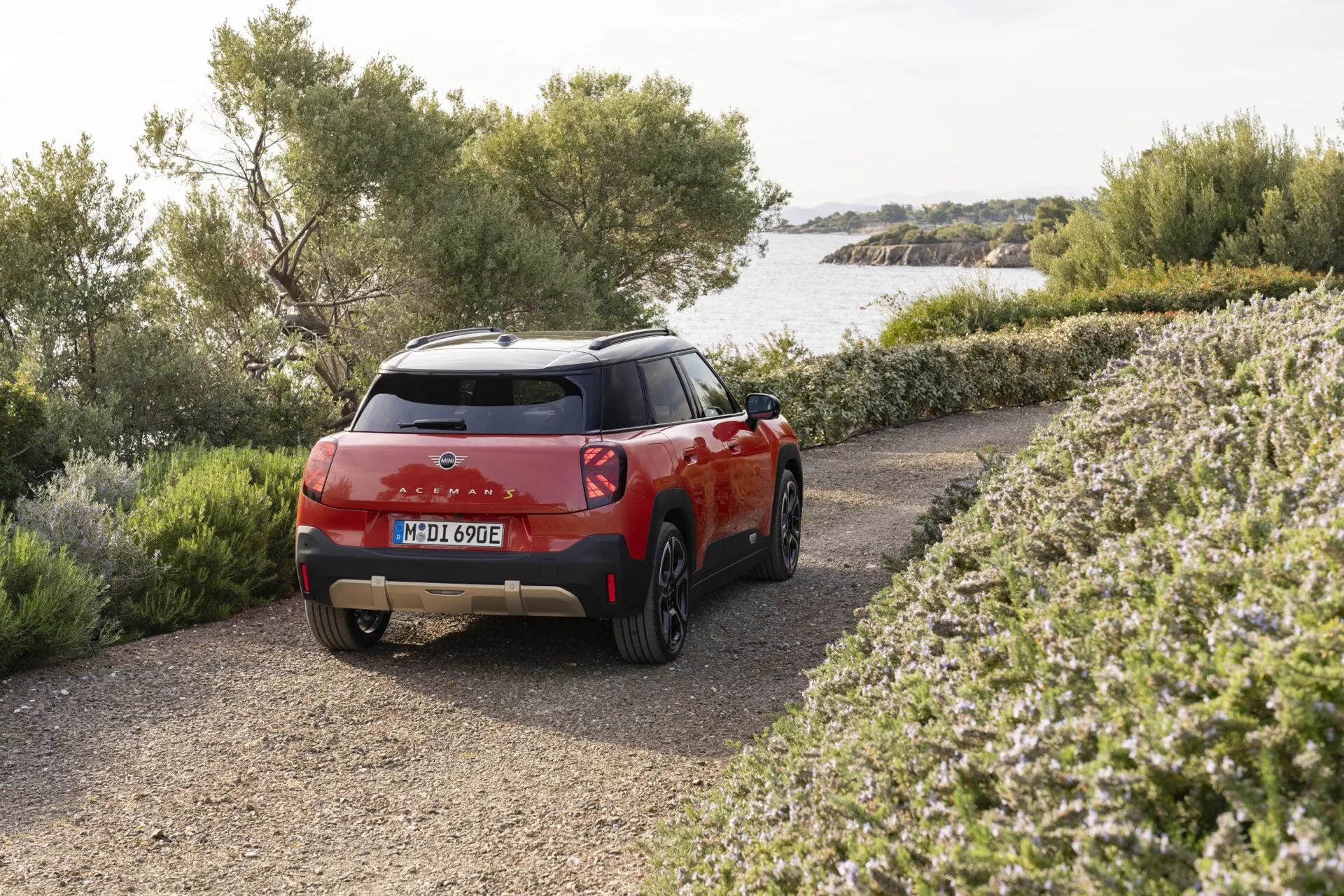 SUV compact Mini Aceman 2024 en rouge garé en extérieur présentant une vue arrière trois quarts soulignant les feux arrière LED, le schéma de couleurs bicolore et les caractéristiques du modèle électrique ou hybride écologique contre un arrière-plan rural ou côtier avec des arbustes et de l'eau