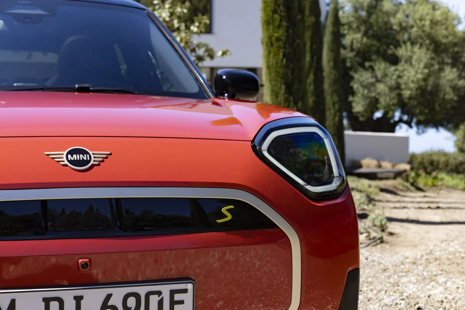 Vue rapprochée d'une Mini Aceman rouge (J05) de 2024 présentant le phare LED élégant, la grille sportive avec badge 'S' et l'emblème électrique 'E' sur le côté avant gauche, reflétant son héritage hybride électrique sportif et design luxueux sous une lumière extérieure éclatante.