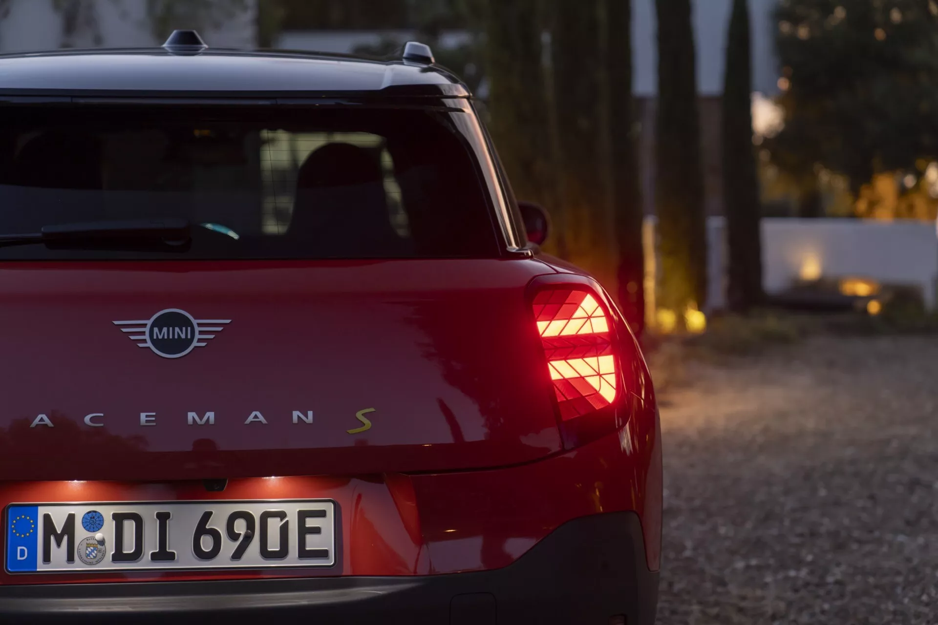 Vue arrière partielle d'une MINI Cooper Paceman S rouge au crépuscule, présentant sa bande verticale de feu arrière, peinture brillante et inscription du modèle, sur fond bokeh sur un chemin non pavé.