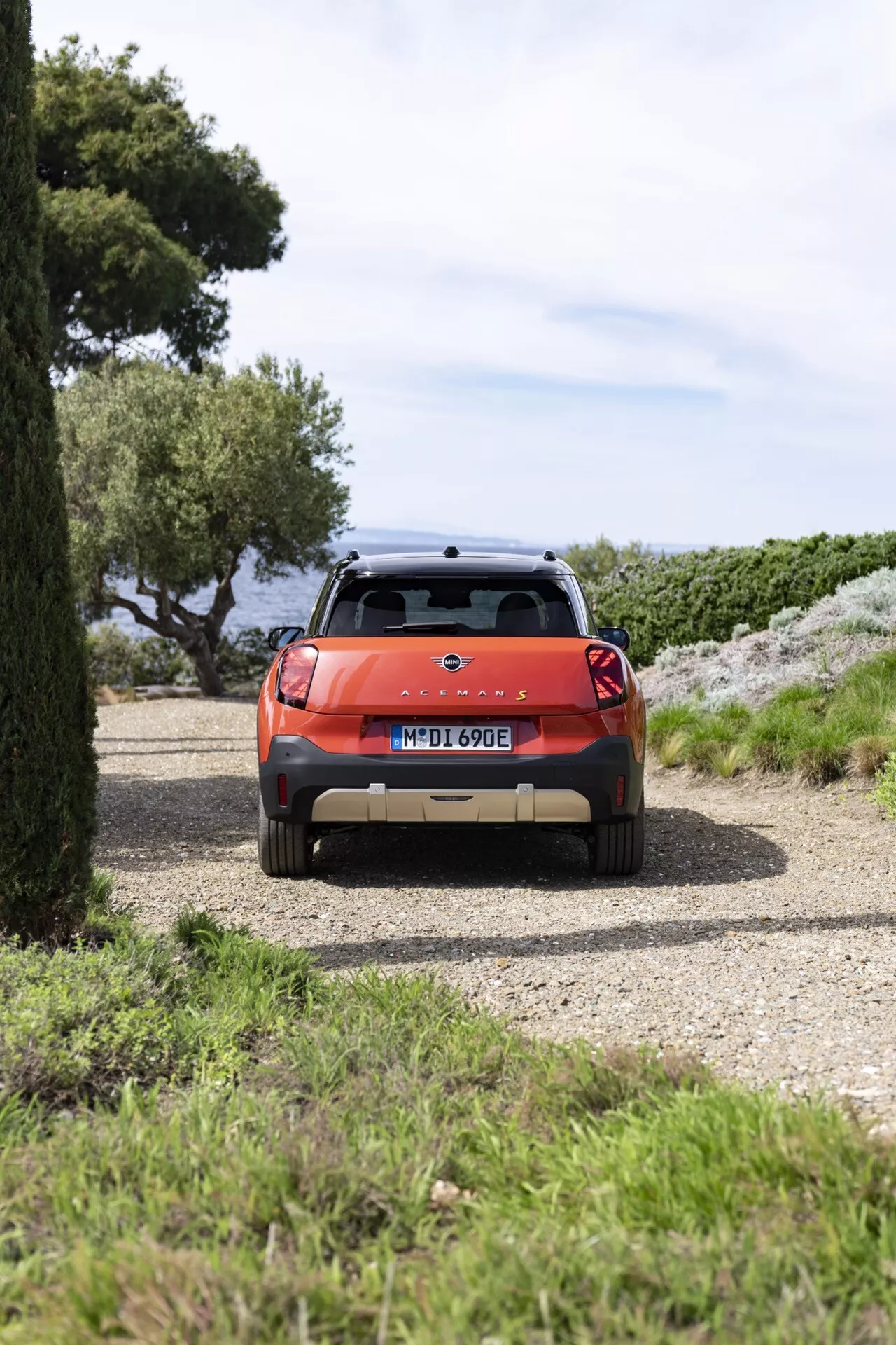 SUV Mini Aceman (J05) 2024 en orange métallique vif, avec feux arrière, essuie-glace arrière, plaque d'immatriculation, aileron arrière avec feu de stop, barres de toit pour chargement supplémentaire, garé sur une allée de gravier avec des arbres et un arrière-plan d'océan possible, mettant en avant son potentiel pour l'aventure et les activités en plein air