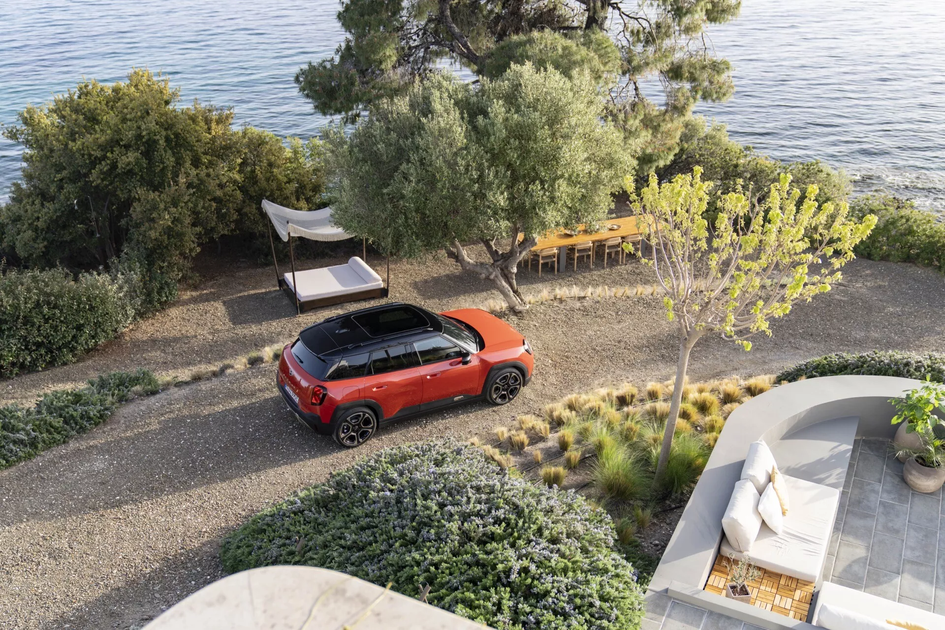 Vue aérienne d'une Mini Aceman 2024 (J05) en rouge vif avec un toit sombre contrasté garée sur une allée de galets dans un cadre naturel près d'une côte, montrant son design moderne de crossover et laissant entrevoir des technologies automobiles avancées.