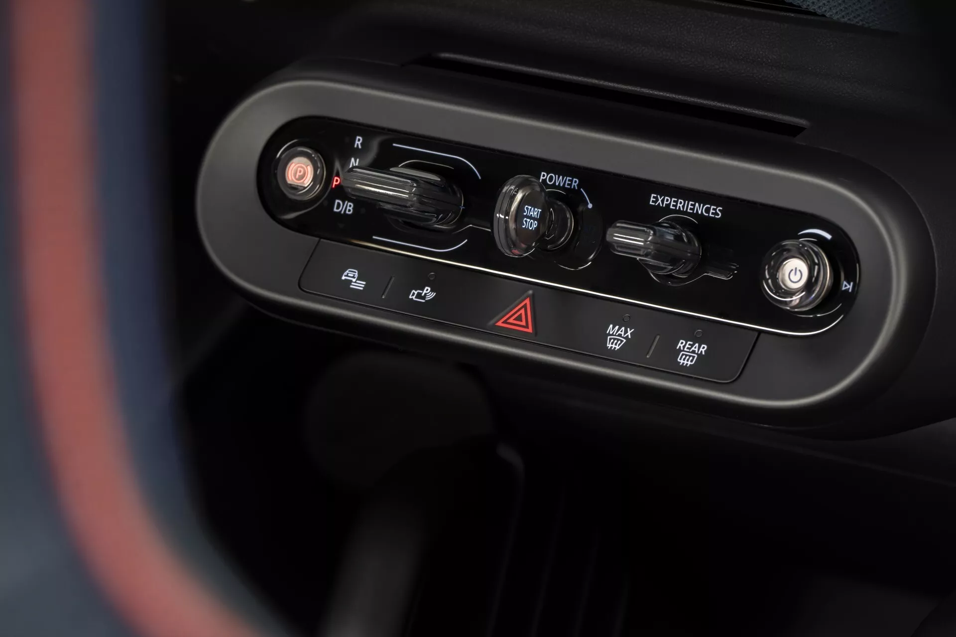 Vue rapprochée du panneau de contrôle de la console centrale du Mini Aceman (J05) 2024 du point de vue du conducteur, présentant des boutons rotatifs et des boutons pour les fonctions du véhicule, y compris l'ajustement du rétroviseur, l'allumage, la sélection des expériences de conduite, les feux de détresse, le dégivrage, le chauffage de la lunette arrière et le volant chauffant, avec une finition chromée haut de gamme