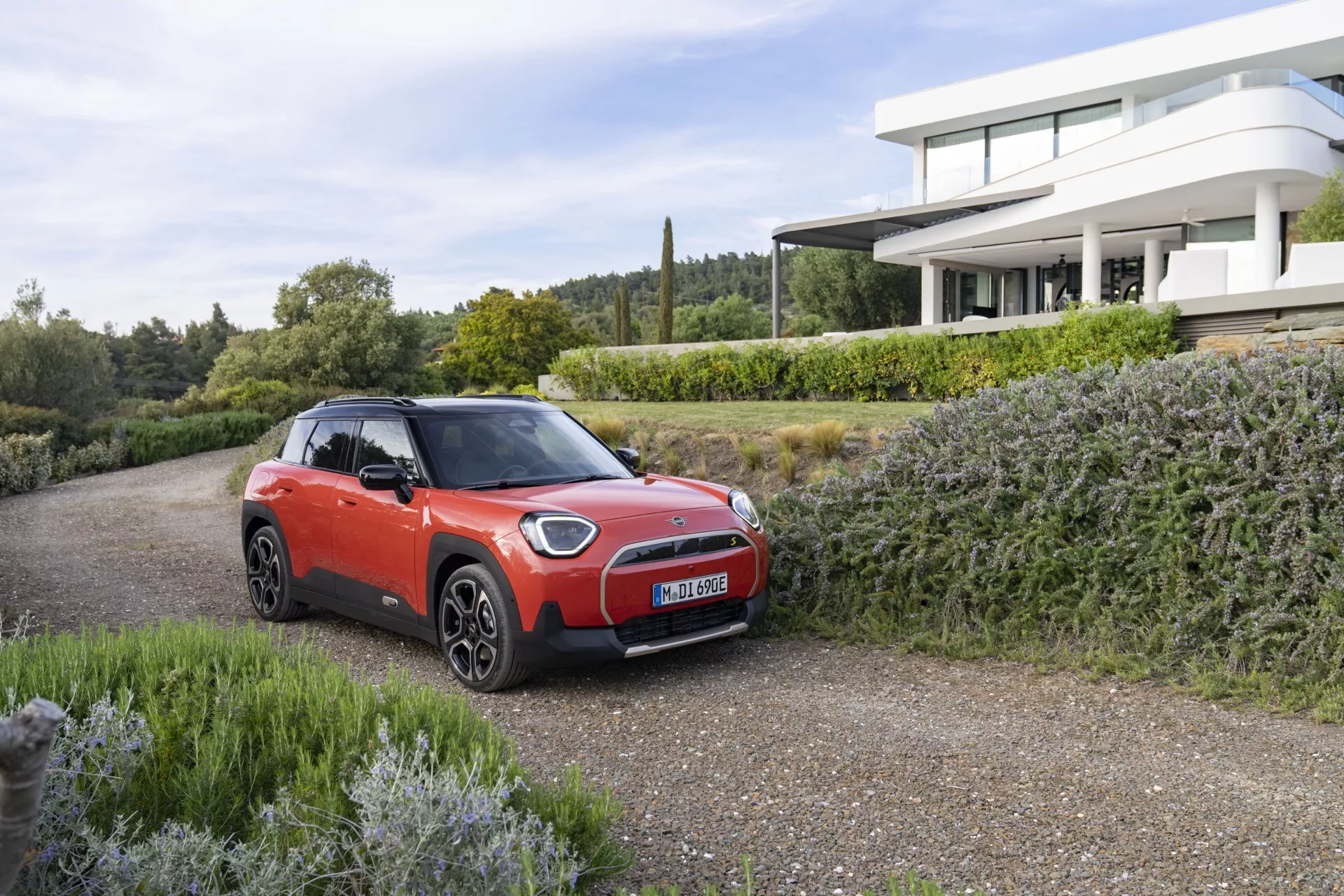 SUV sous-compact Mini Aceman 2024 en rouge avec toit noir, mettant en avant des phares LED modernes et des jantes alliage aérodynamiques garé sur une allée de gravier avec verdure et maison haut de gamme en arrière-plan