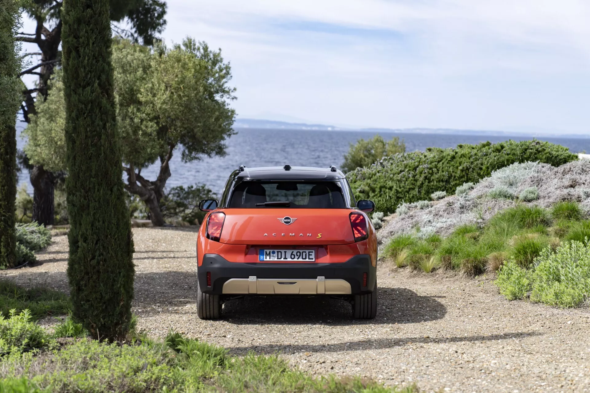 Vue arrière du Mini Aceman (J05) 2024 en orange, avec un spoiler sportif et des feux arrière LED, situé sur un chemin de gravier au bord de la mer avec un paysage naturel en arrière-plan en journée