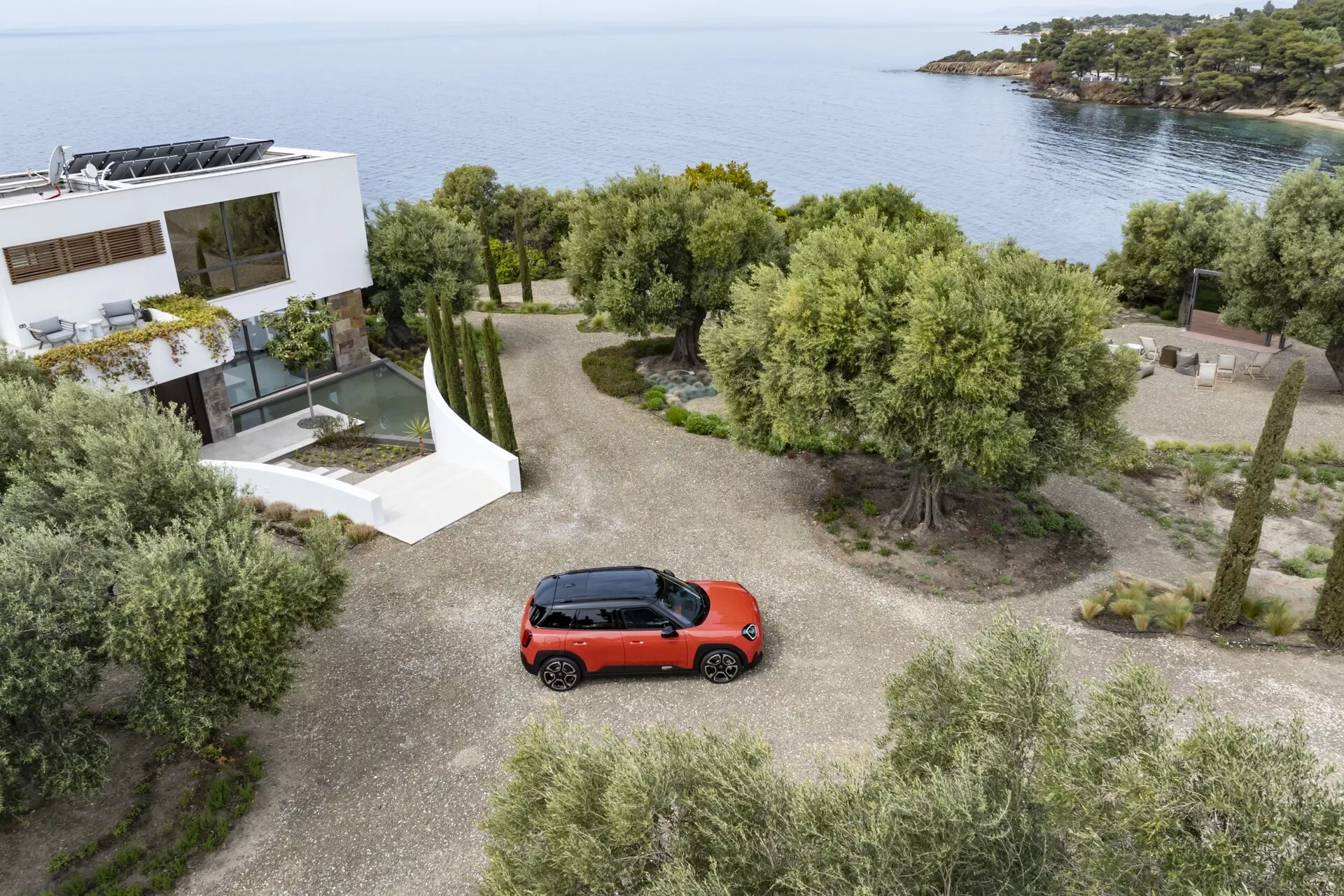 Mini Aceman (J05) hatchback rouge de 2024 garée sur une allée en gravier avec vue aérienne montrant entièrement le dessus du véhicule, dans un paysage pittoresque avec des arbres, des arbustes et une vue sur la mer à côté d'une maison moderne