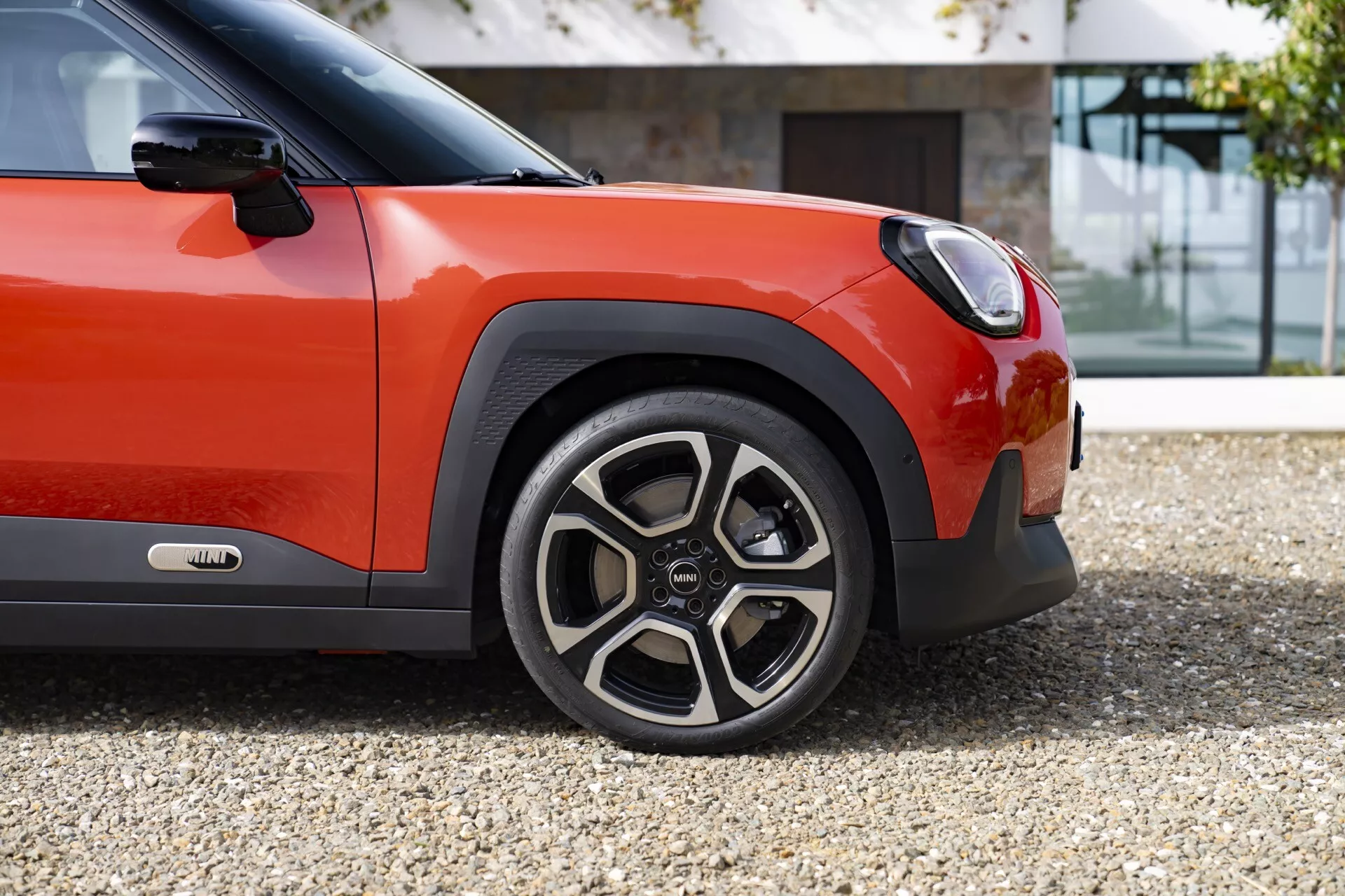 Mini Aceman 2024 (J05) présentant une vue de côté rapprochée de la roue avant sportive avec un design à rayons multiples distinctif et jante bicolore, complétée par la carrosserie bicolore rouge orangé vif et noir du quart avant du véhicule, et un aperçu de la technologie avancée des phares LED
