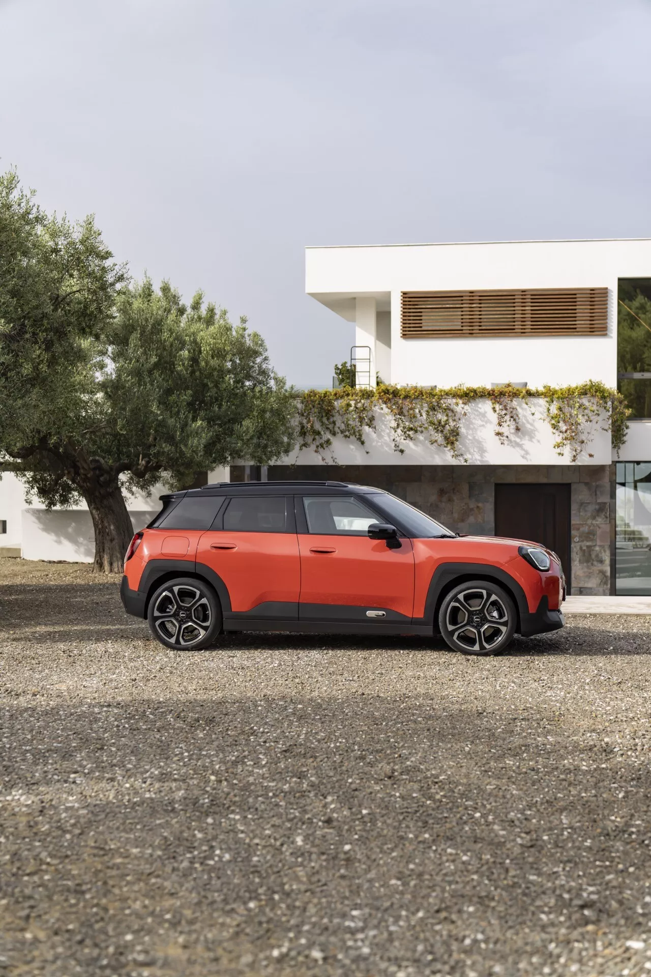 Mini Aceman 2024 (J05) garée sur le gravier à l'extérieur d'une maison moderne, affichant son profil latéral avec une vue de trois quarts présentant un schéma de couleurs bicolores, des poignées de porte affleurantes et des roues à multiples branches
