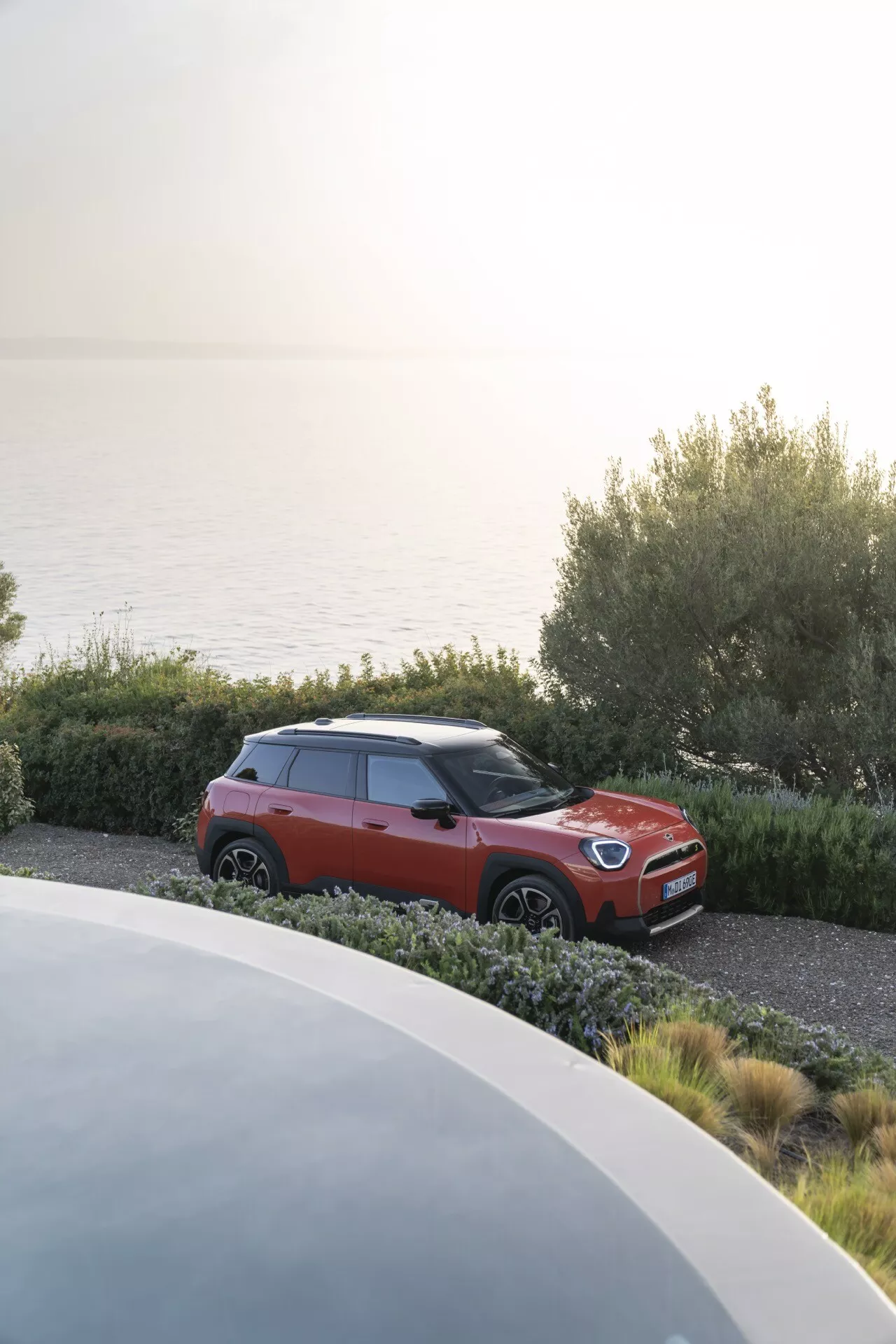 Mini Aceman (J05) de 2024 garée dans un paysage côtier pittoresque avec carrosserie bicolore rouge et noir, équipée de phares LED, de jantes en alliage, de rétroviseurs avec clignotants intégrés, de vitres teintées et de barres de toit, vue sous un angle élevé avant gauche