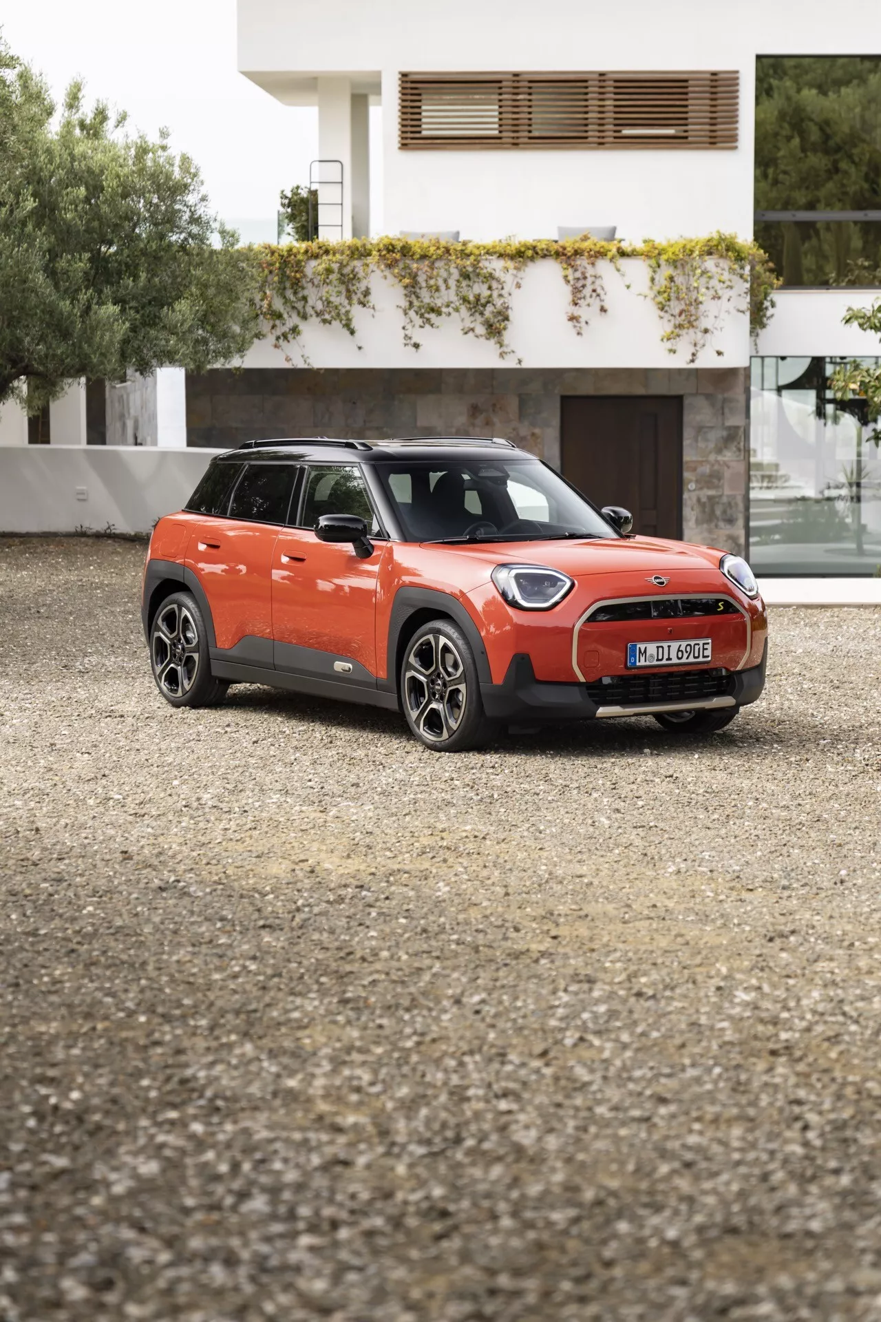 SUV compact Mini Aceman (J05) 2024 en orange vibrant avec toit noir, présenté sous un angle avant quart sur une surface en gravier, mettant en vedette des phares LED modernes, des roues en alliage et un design de grille aérodynamique sur fond résidentiel.