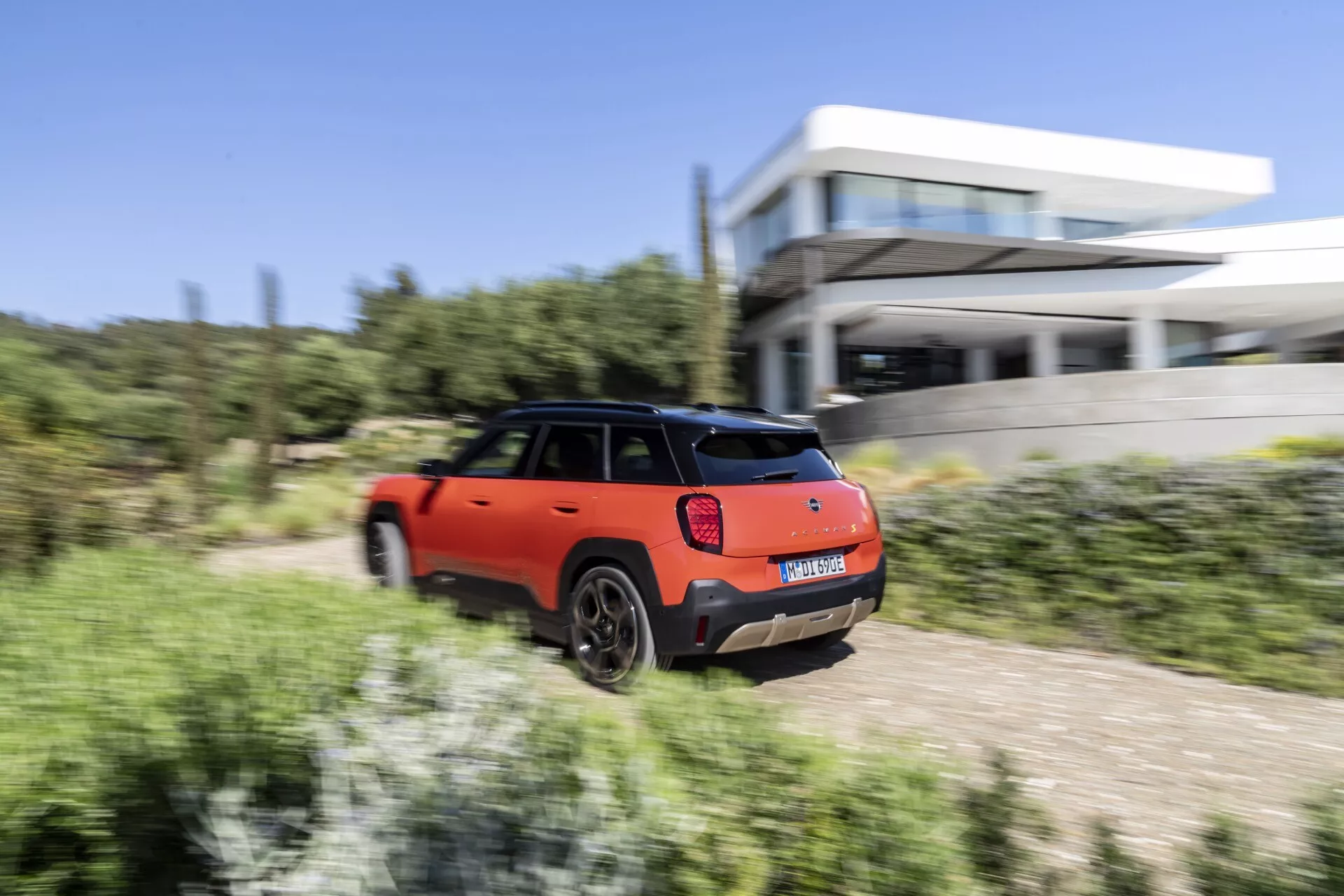 Mini Aceman (J05) 2024 en mouvement, vue arrière et de côté mettant en évidence sa peinture bicolore orange et noire, feux arrière LED et esthétique sportive sur un fond dynamique comprenant végétation naturelle et architecture moderne