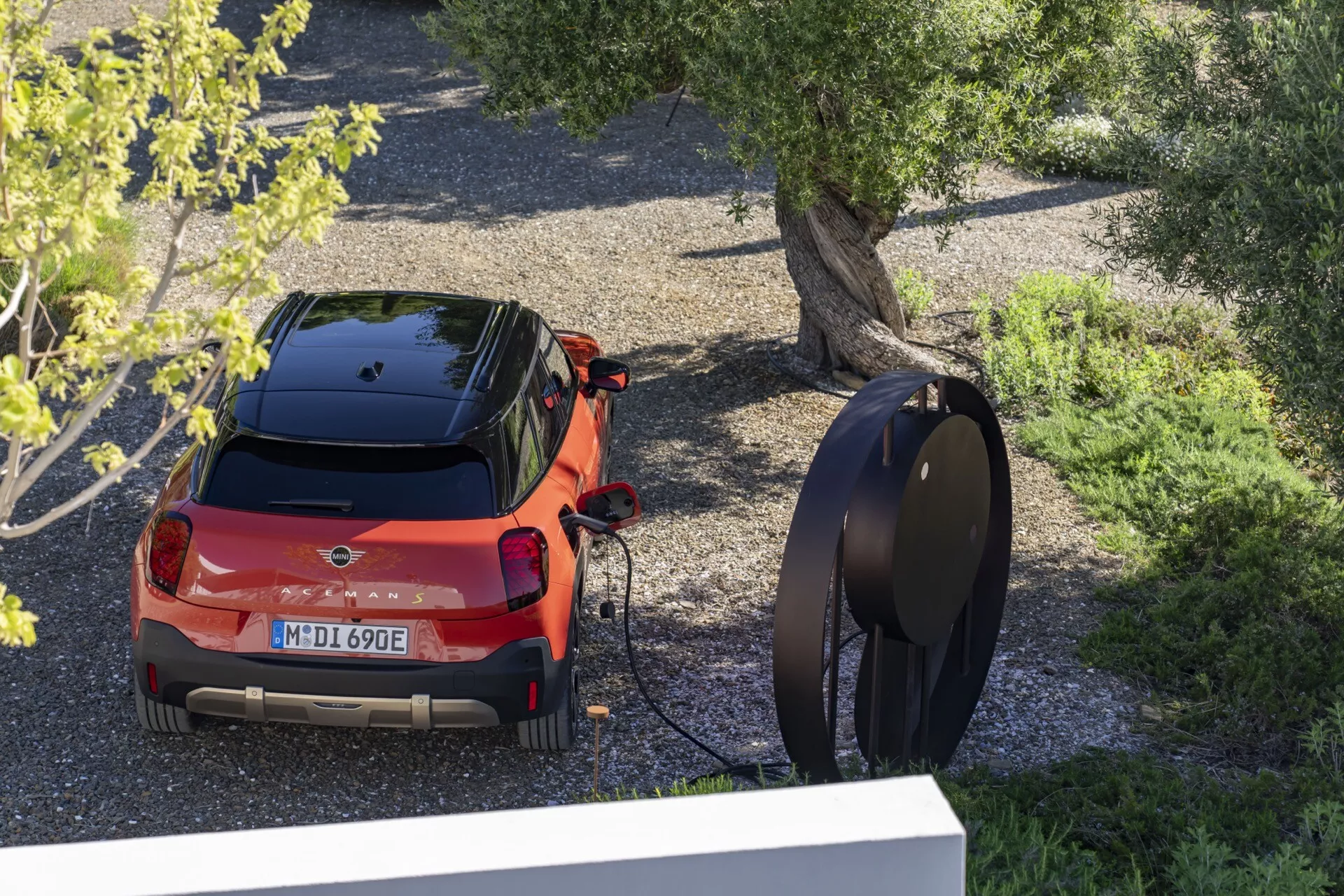 Vue arrière en plongée d'une voiture électrique Mini Aceman (J05) hatchback rouge de 2024 branchée à une station de recharge moderne, garée en extérieur entourée d'arbres et de gravier.
