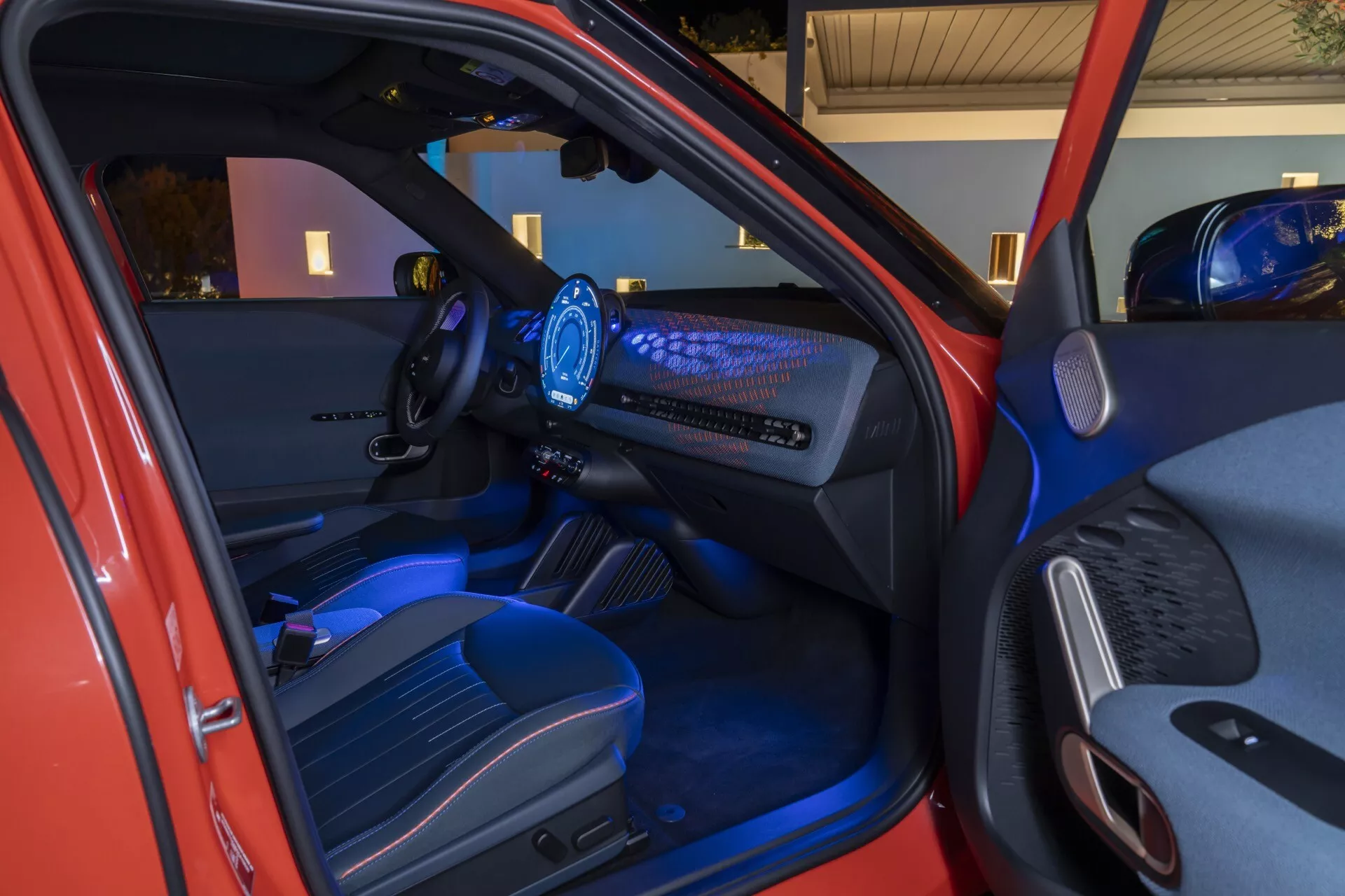 Vue intérieure du Mini Aceman 2024 (JO5) montrant la porte côté conducteur, volant ergonomique, tableau de bord numérique, et la zone du passager avant avec éclairage d'ambiance bleu et fonctionnalités technologiques avancées