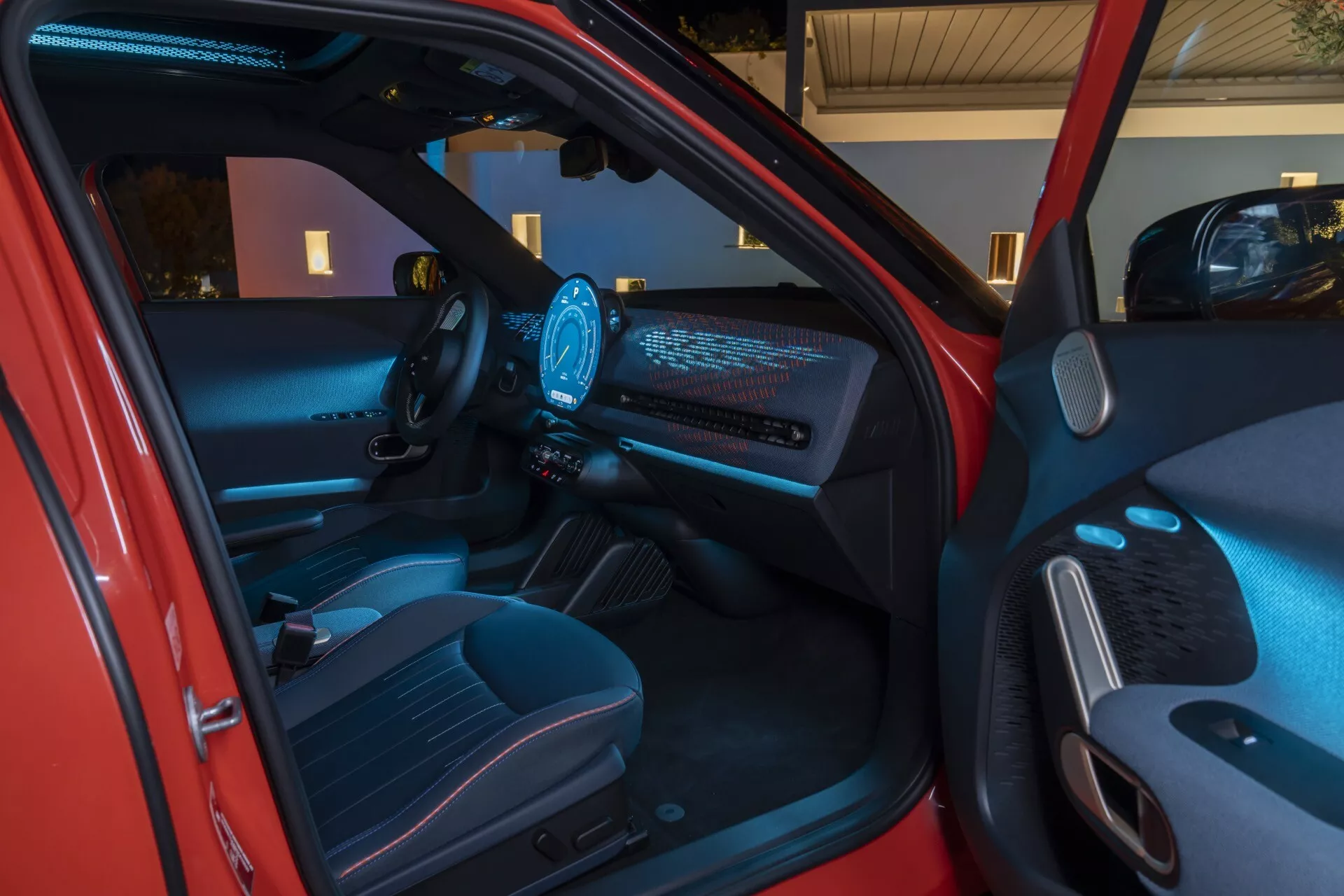 Vue intérieure du Mini Aceman 2024 (J05) montrant un tableau de bord futuriste avec écran illuminé, volant moderne avec commandes illuminées, cluster d'instruments numériques et éclairage d'ambiance high-tech depuis un angle extérieur à proximité du côté conducteur.