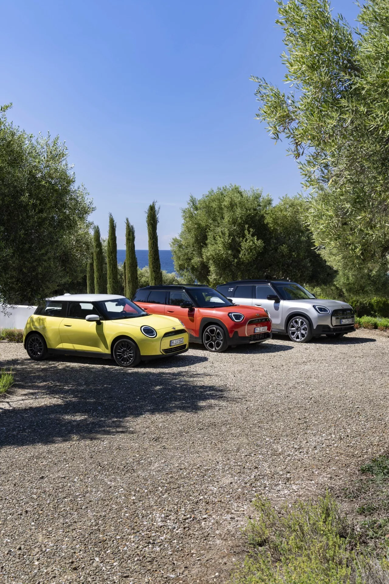 Gamme Mini Aceman 2024 présentant une hatchback compacte jaune vif, un modèle compact rouge vibrant et un SUV de couleur métallique claire garés côte à côte sur gravier avec des feuillages verts et un ciel bleu clair en arrière-plan, mettant en lumière des designs modernes et des caractéristiques technologiques