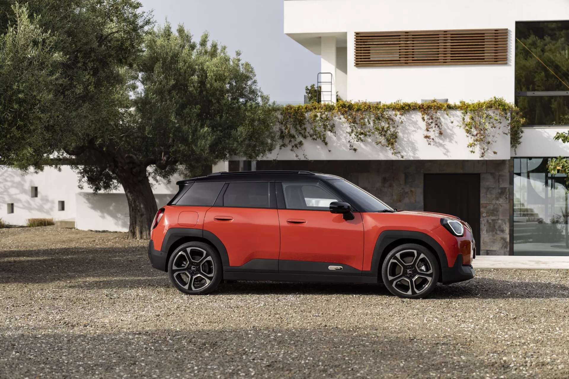 SUV compact Mini Aceman 2024 bicolore rouge et noir garé à l'extérieur devant un bâtiment résidentiel moderne, présentant son profil latéral avec des détails du véhicule et des éléments de design, incluant des roues en alliage à plusieurs branches, toit panoramique et technologies de sécurité avancées.