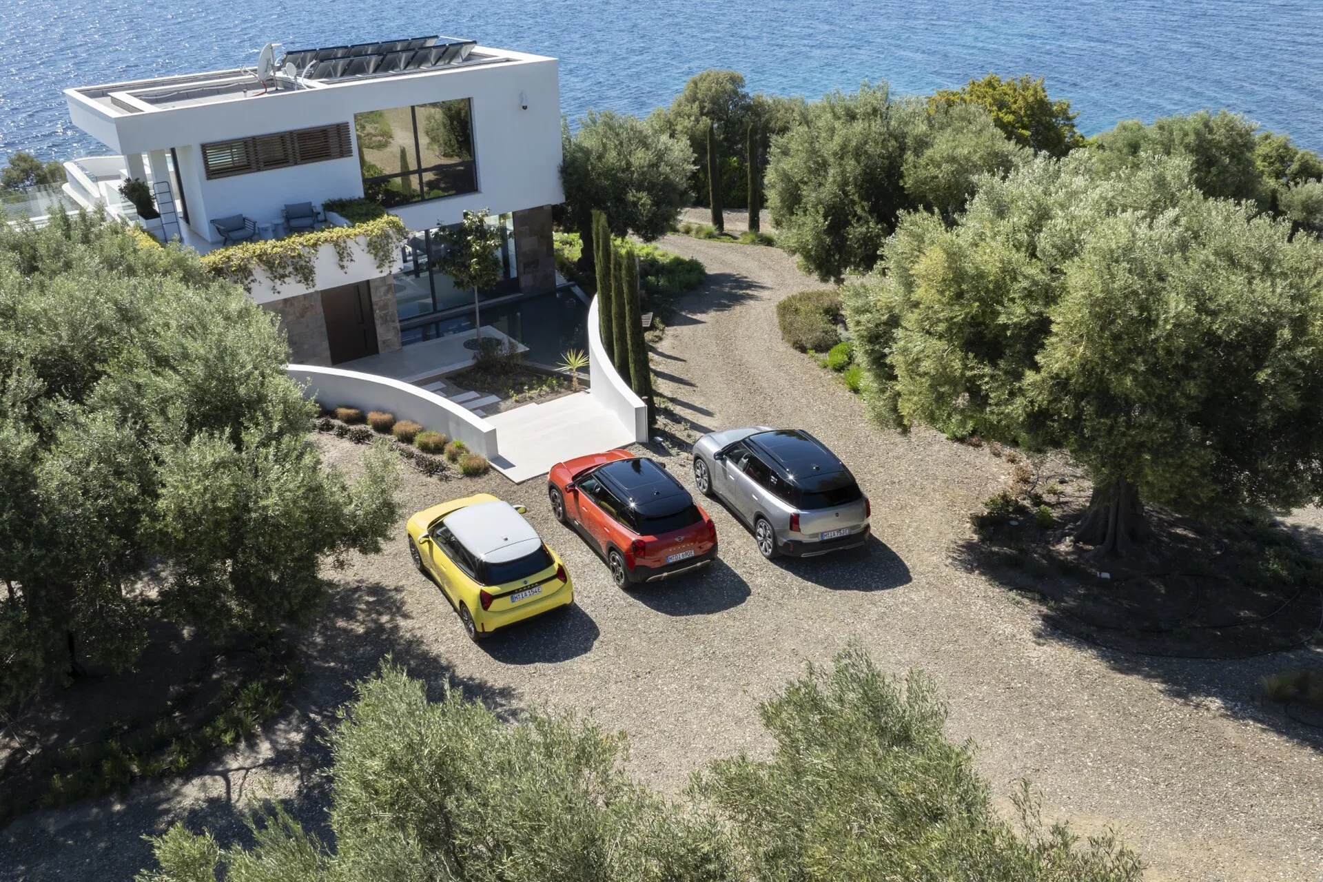 Vue aérienne d'une hatchback jaune, d'une berline gris métallisé et d'un crossover rouge garés à l'extérieur d'une maison contemporaine au milieu de la végétation méditerranéenne