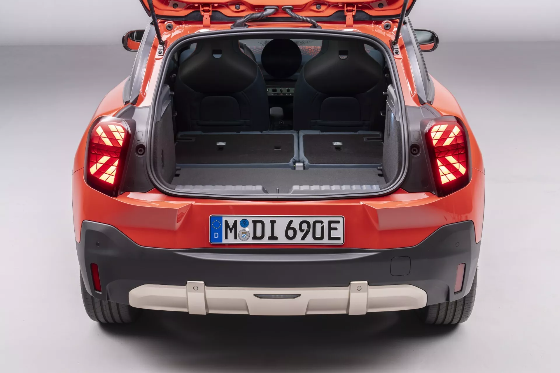 Vue arrière d'une Mini Aceman 2024 (J05) orange-rouge avec hayon ouvert, montrant l'espace de chargement vide, feux arrière LED avancés, capteurs d'aide au stationnement, caméra de recul et indication potentielle de véhicule électrique à partir de la plaque d'immatriculation de style européen, sur un fond de studio
