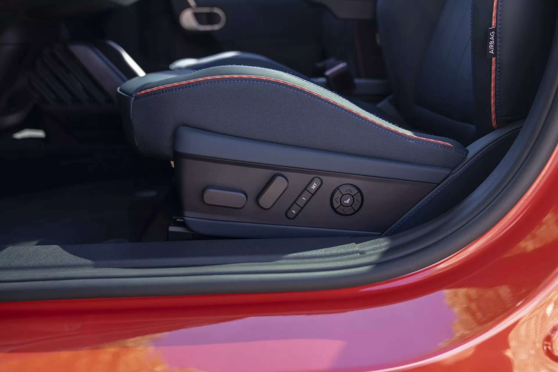 Vue détaillée de l'intérieur de la MINI Aceman (J05) 2024 montrant le siège conducteur avec commandes de réglage électroniques et fonction mémoire, design sportif avec soutien latéral et coutures contrastées, extérieur rouge à travers la porte ouverte