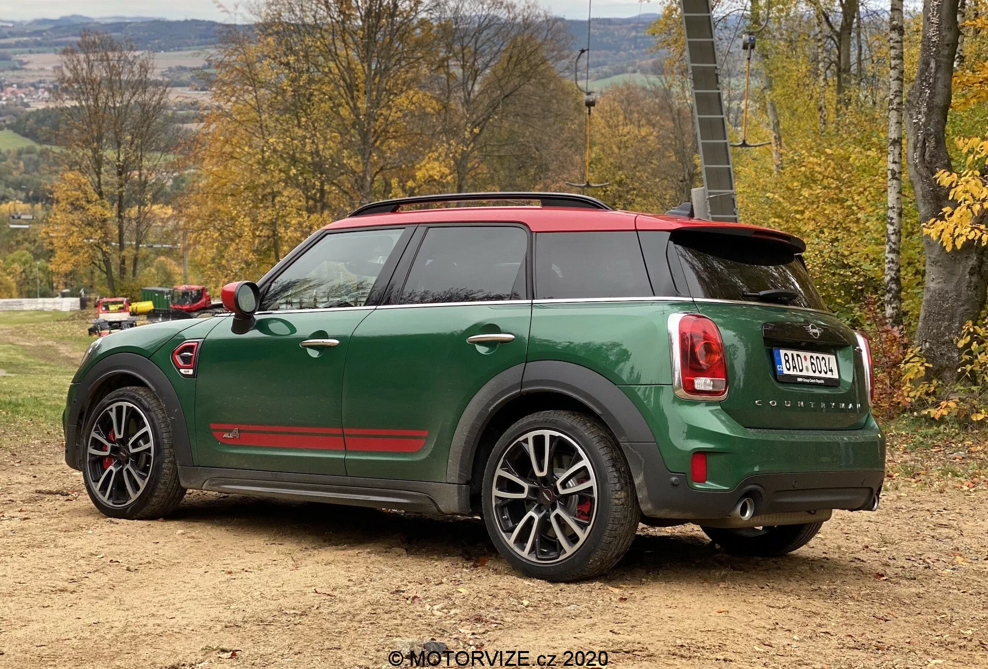 Vue arrière trois-quarts du Mini Cooper Countryman vert 2020 en édition sportive John Cooper Works, avec toit rouge et coques de rétroviseurs, double sortie d'échappement, aileron arrière, jantes en alliage à cinq branches avec détails noirs et fonctionnalités technologiques avancées comme des capteurs de stationnement et une antenne aileron de requin, positionné sur une surface en terre dans un environnement rural d'automne.