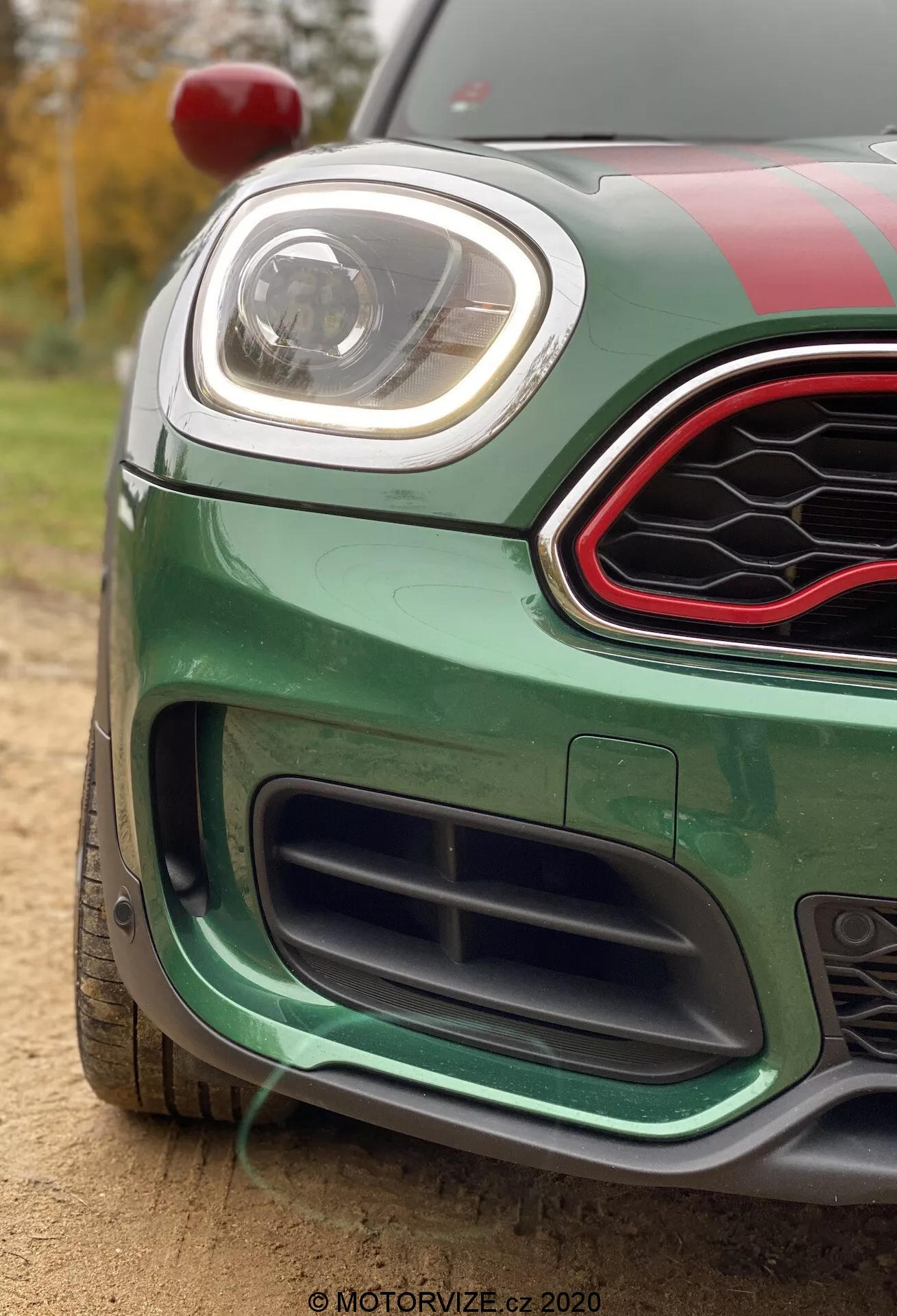 Image en gros plan d'une Mini Countryman John Cooper Works 2020 verte vibrante, centrée sur le quart avant-gauche. Capturée à un angle bas, l'image met en évidence la grille inférieure avec un design en nid d'abeille noir, le pare-chocs avant avec des caractéristiques aérodynamiques et le phare LED élégant. La grille supérieure est hexagonale avec une finition noire brillante et une bordure accent rouge. La voiture présente des bandes de course sur le capot et le toit, un rétroviseur latéral rouge visible en haut à gauche et se trouve à l'extérieur avec une lumière naturelle et des arbres et feuillage en arrière-plan flou.