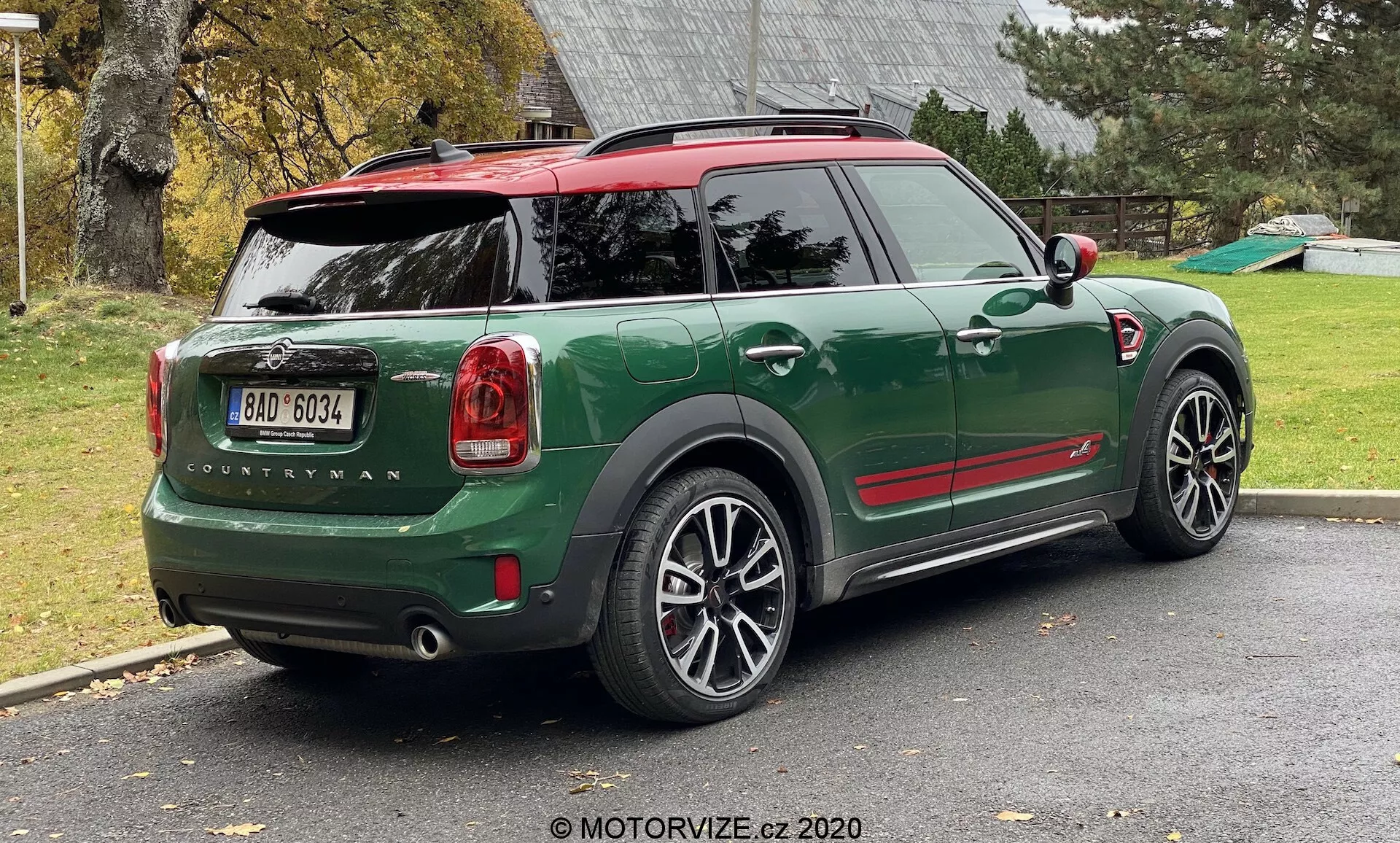 Vue arrière trois quarts d'un MINI Countryman John Cooper Works 2020 vert garé dans une allée résidentielle. L'image montre toute la moitié arrière du côté droit de la voiture, y compris le hayon arrière, la roue arrière droite, une partie du toit et les portes passagers. Le design sportif et compact du SUV présente des accents rouges contrastants sur les coques des rétroviseurs latéraux et le toit, des arches de roue noires et de grandes jantes en alliage multi-rayons. Les feux arrière, le nom du modèle 'Countryman' et le logo MINI sont clairement visibles. La voiture a un toit rouge avec un spoiler et des rails de toit, et la lunette arrière comprend un dégivreur et un essuie-glace.
