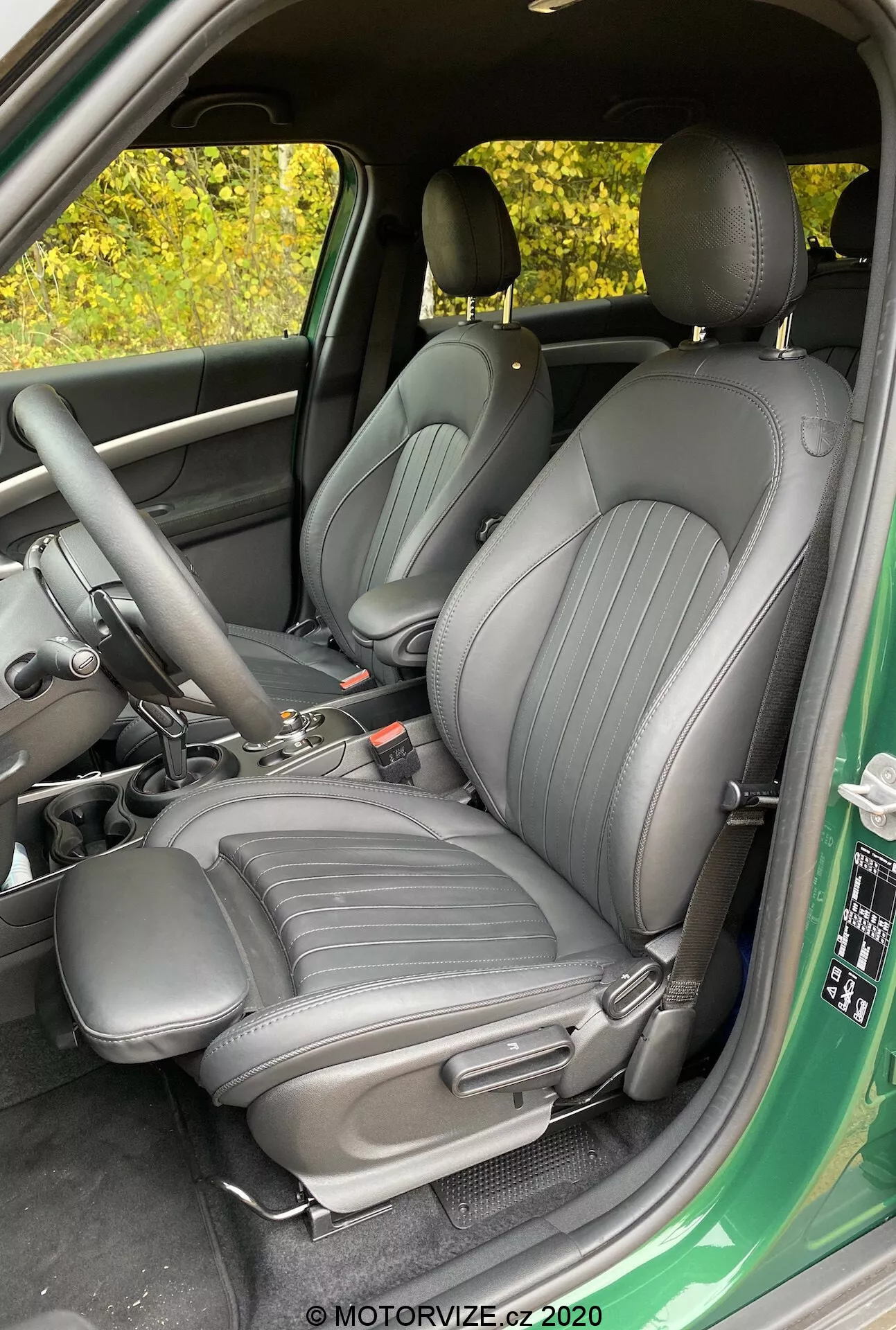 Image de l'intérieur de la Mini Countryman John Cooper Works 2020 se concentrant sur les sièges avant du conducteur et du passager, rembourrés en matériau de haute qualité foncé avec des lignes verticales, vue depuis la porte du conducteur ouverte, montrant la console centrale avec le levier de vitesse et le frein à main, vue partielle du panneau de porte avec les commandes pour les fenêtres et les rétroviseurs.