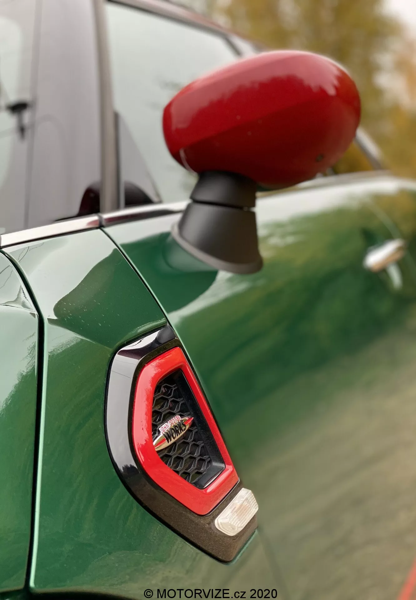 Gros plan sur le rétroviseur latéral rouge vif et l'indicateur latéral du côté conducteur d'une Mini Countryman John Cooper Works 2020, vu contre un panneau de carrosserie vert métallisé profond avec des reflets d'un ciel couvert et des arbres. La mise au point met en valeur l'indicateur angulaire avec une lentille transparente montrant probablement une technologie LED, entouré d'une finition sportive et brillante.