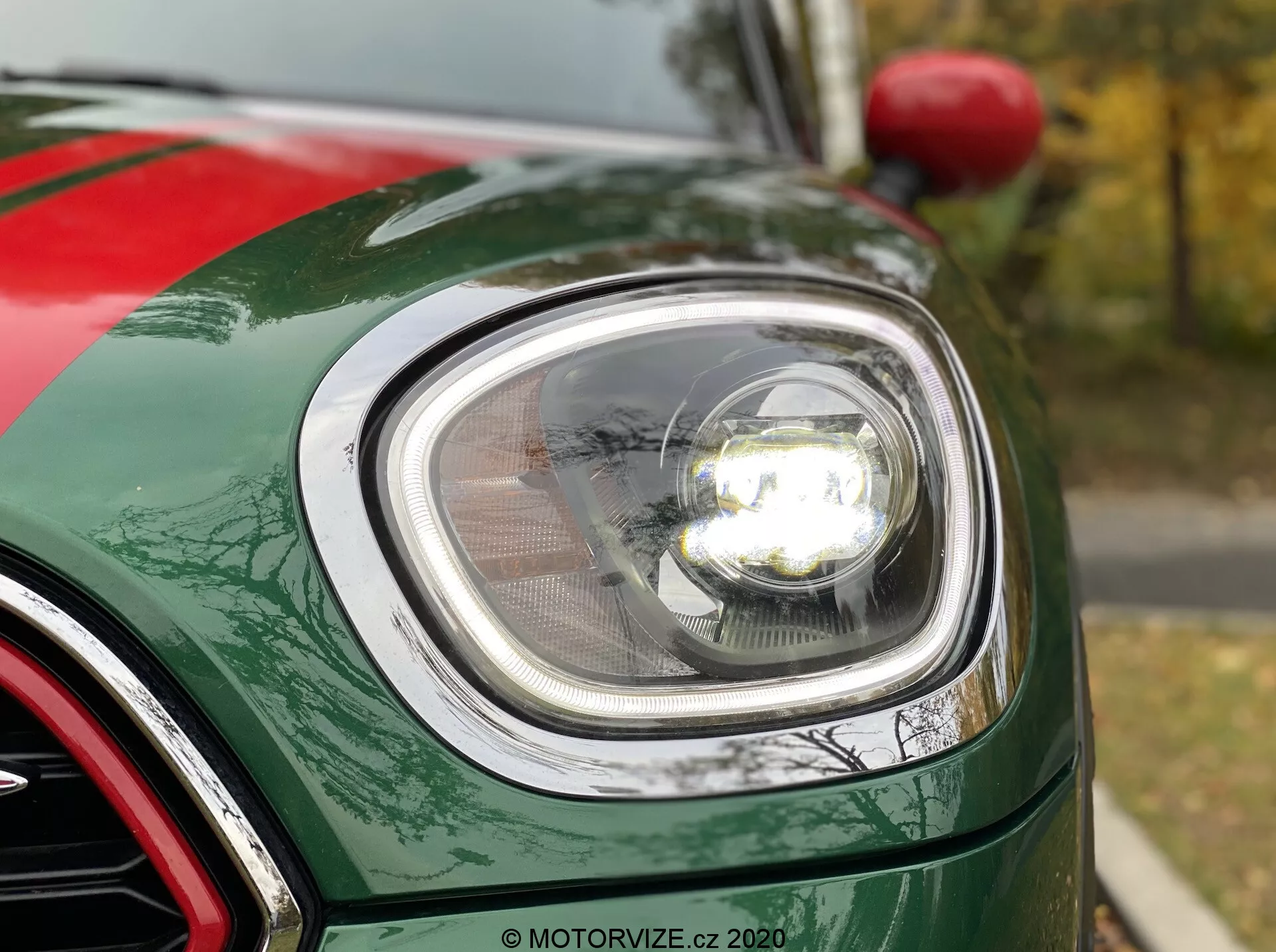 Vue rapprochée de la partie avant gauche d'une Mini Countryman John Cooper Works 2020, concentrée sur le phare rond moderne avec technologie LED, couvercle de lentille clair et projecteur éclairé. L'image montre la courbure du garde-boue avant, le bord du capot et une partie de la calandre à finition chromée. La voiture présente une peinture verte métallique brillante avec des paillettes métalliques visibles.