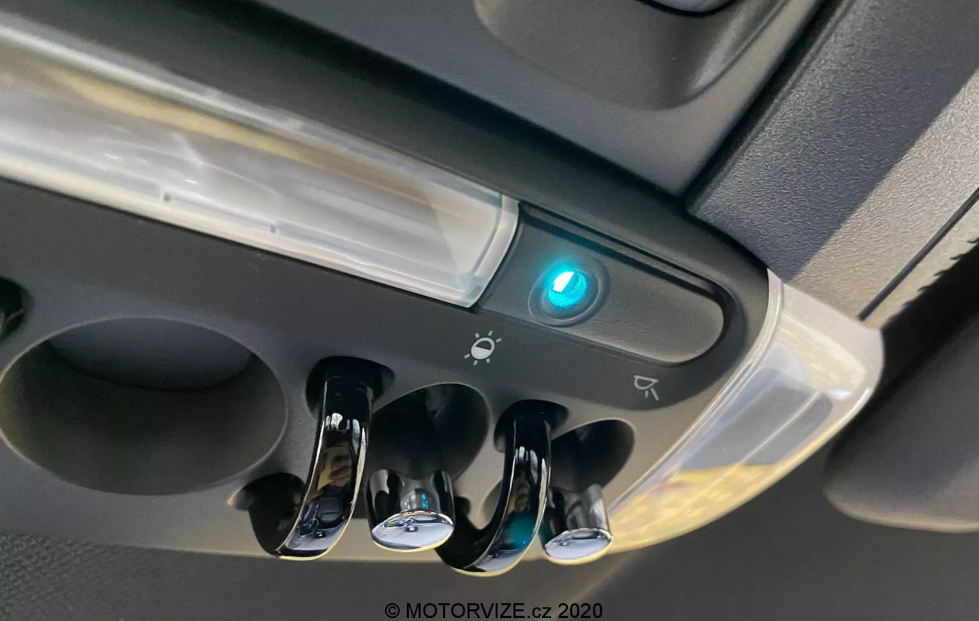 Vue rapprochée de la console supérieure du Mini Countryman 2020 John Cooper Works montrant les lumières intérieures de l'habitacle, trois interrupteurs à bascule chromés pour les lumières individuelles de lecture, une lumière centrale d'habitacle, un bouton éclairé en bleu pouvant indiquer la connectivité Bluetooth ou une fonction de sécurité, le bord d'un possible compartiment de rangement, des icônes suggérant des commandes ou des capteurs supplémentaires, et un design moderne avec des finitions brillantes et mates.