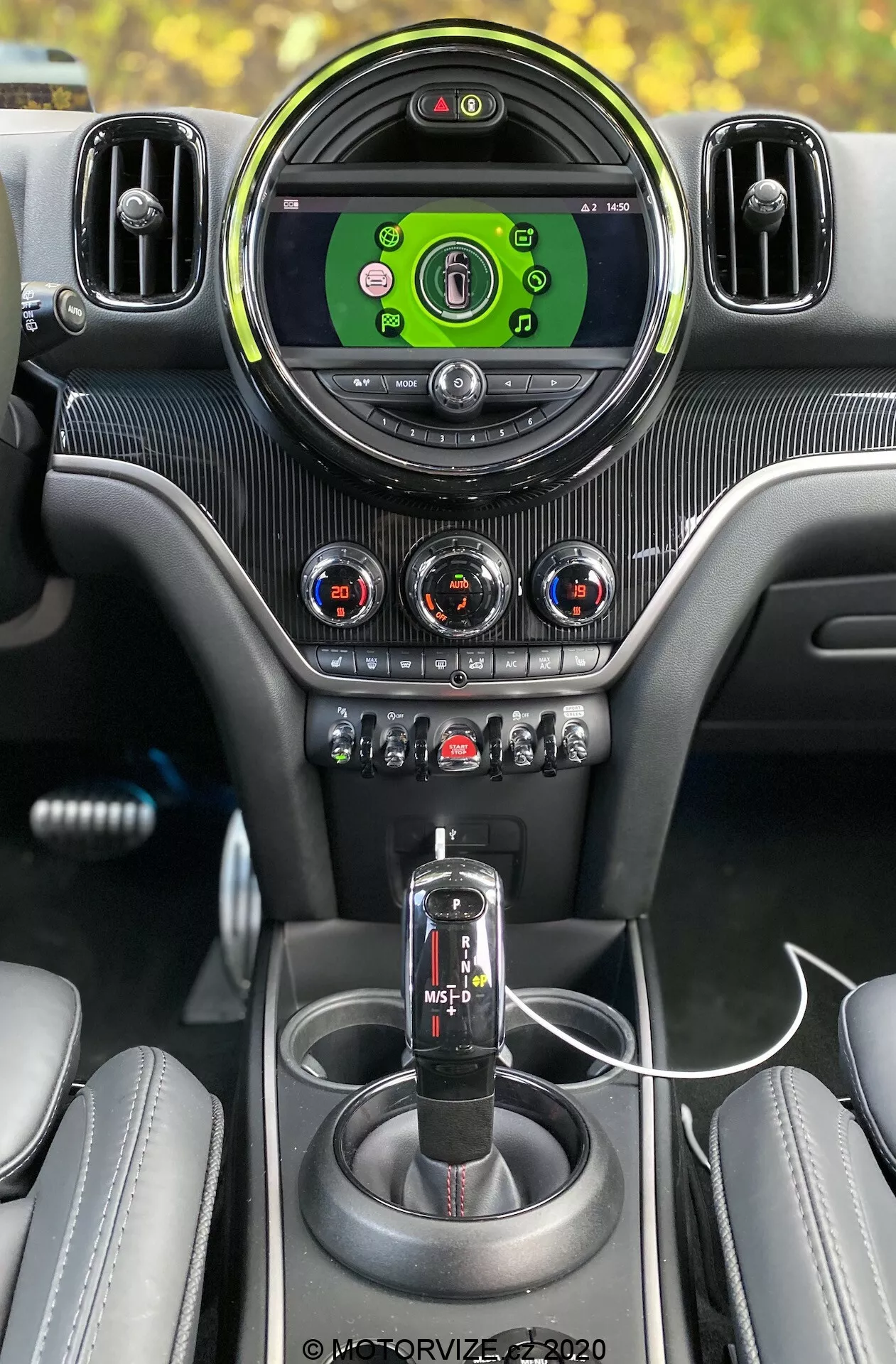 Vue_intérieure_d'une_Mini_Countryman_John_Cooper_Works_2020_depuis_la_perspective_du_conducteur,_soulignant_les_caractéristiques_de_premium_comme_l'écran_circulaire_central_du_système_d'infodivertissement_avec_une_interface_verte,_encadrées_par_des_aérateurs_ajustables_au_contour_argenté;_trois_commandes_rotatives_de_climatisation_avec_indicateurs_rouges_et_bleus_dans_un_panneau_à_motif_de_fibre_de_carbone;_une_range_de_boutons_et_d'interrupteurs_pour_diverses_fonctions_de_la_voiture,_y_compris_un_mode_sport/traction;_un_levier_de_vitesse_automatique_noir_avec_une_option_de_mode_manuel;_une_console_centrale_cohésive_avec_motif_en_fibre_de_carbone;_deux_portes-gobelets;_un_câble_de_chargement_USB_connecté;_et_des_sièges_partiellement_visibles_en_cuir_cousu,_tous_les_éléments_combinaient_pour_créer_un_intérieur_automobile_premium_et_technologiquement_orienté.