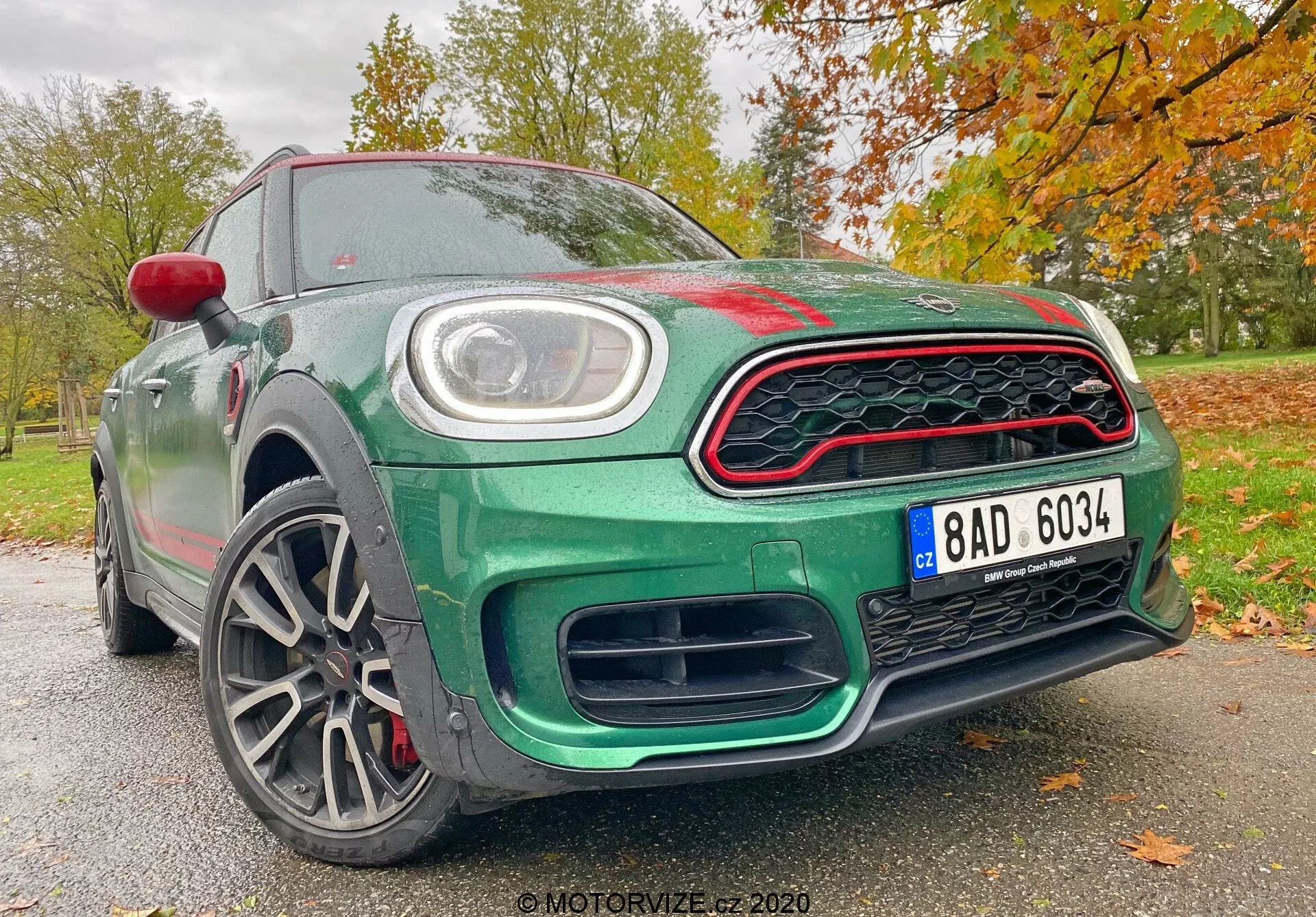 L'image présente la Mini Countryman 2020 édition John Cooper Works sous un angle avant bas, mettant en valeur son design sportif et moderne. Parmi les éléments visibles, on trouve la calandre avant en forme de nid d'abeille avec des accents rouges, de larges prises d'air sous la calandre pour améliorer le flux d'air, une plaque d'immatriculation centrale et des feux de jour à LED circulaires intégrés dans les phares principaux. Le capot présente une légère bosse avec des plis aérodynamiques, tandis que les lignes sculptées sur le profil latéral renforcent son apparence dynamique. Les jantes en alliage à plusieurs branches suggèrent une construction légère pour une meilleure maniabilité et accélération, et révèlent des disques de frein haute performance. La carrosserie est peinte en vert métallisé, rehaussée par une coque de rétroviseur latéral rouge et des accents rouges sur la calandre, contre un fond automnal de feuilles. Cette perspective souligne les caractéristiques technologiques et de design avancées du véhicule, le rendant remarquable dans les catégories des voitures sportives et de luxe.
