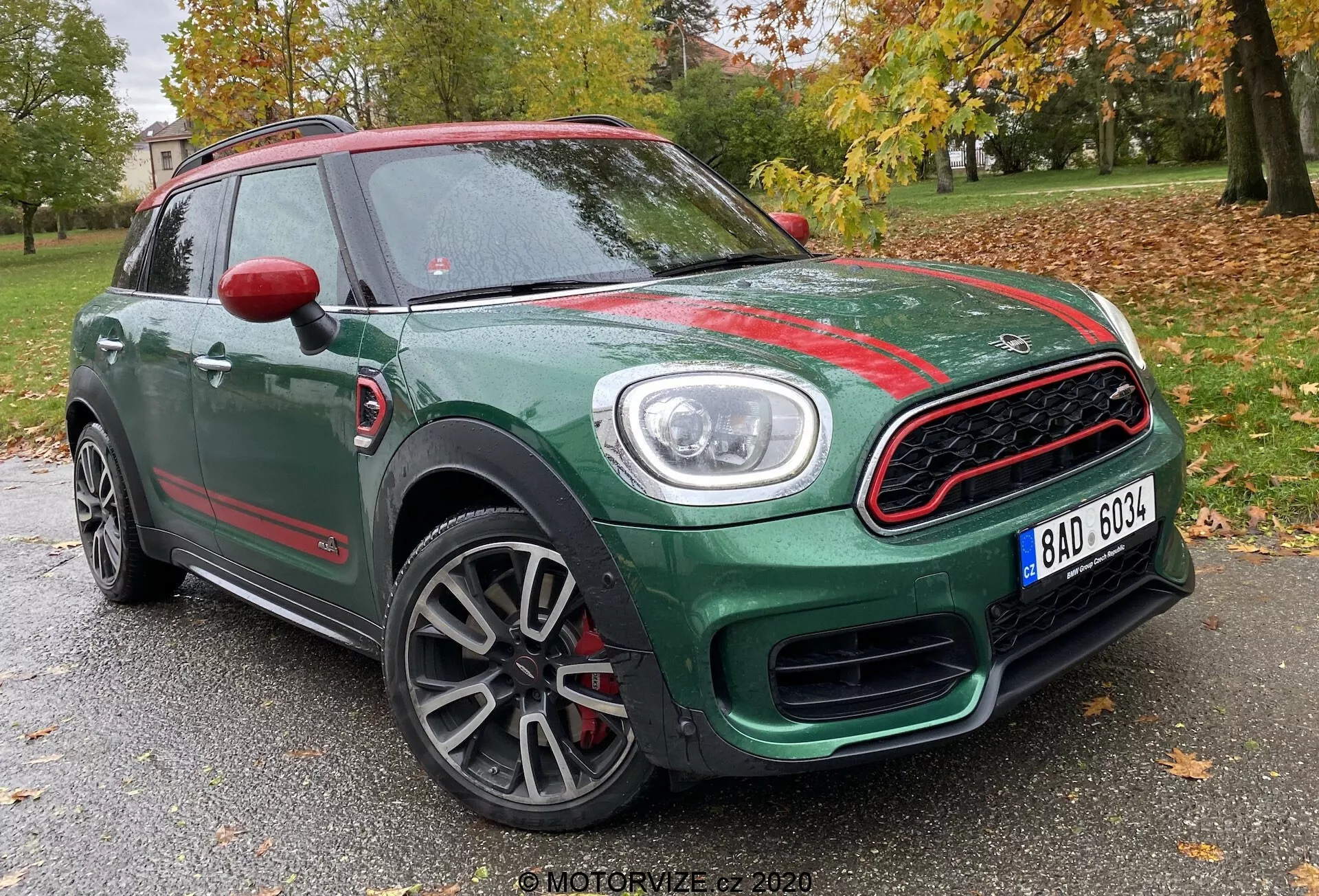 Vue de trois quarts avant d'un Mini Countryman John Cooper Works 2020, montrant sa carrosserie compacte de SUV verte avec des accents rouges contrastants. Capturée sous un angle bas, l'image met en évidence la calandre avant sportive avec un contour chromé et un design en nid d'abeilles, les phares circulaires proéminents avec des feux diurnes LED, les jantes en alliage multibrins avec une finition grise et les capuchons de rétroviseurs rouges. La photo est prise avec un fond d'arbres et une structure faible, suggérant un emplacement dans un parc ou une zone résidentielle.