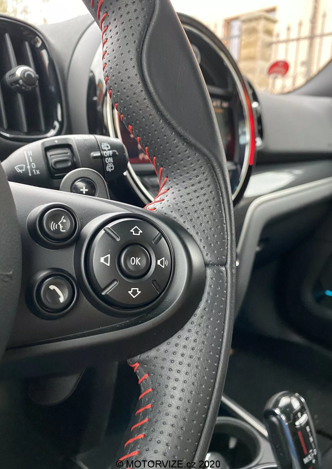 Vue intérieure d'une Mini Countryman John Cooper Works 2020 se concentrant sur le volant en cuir avec des surpiqûres rouges, des boutons de commande pour l'audio et les appels, et un tableau de bord partiellement visible avec des accents rouges suggérant un modèle sportif. Affiche la zone de contrôle du conducteur, le levier de vitesse automatique et les commandes des vitres électriques, mettant en avant la commodité et les fonctionnalités avancées.