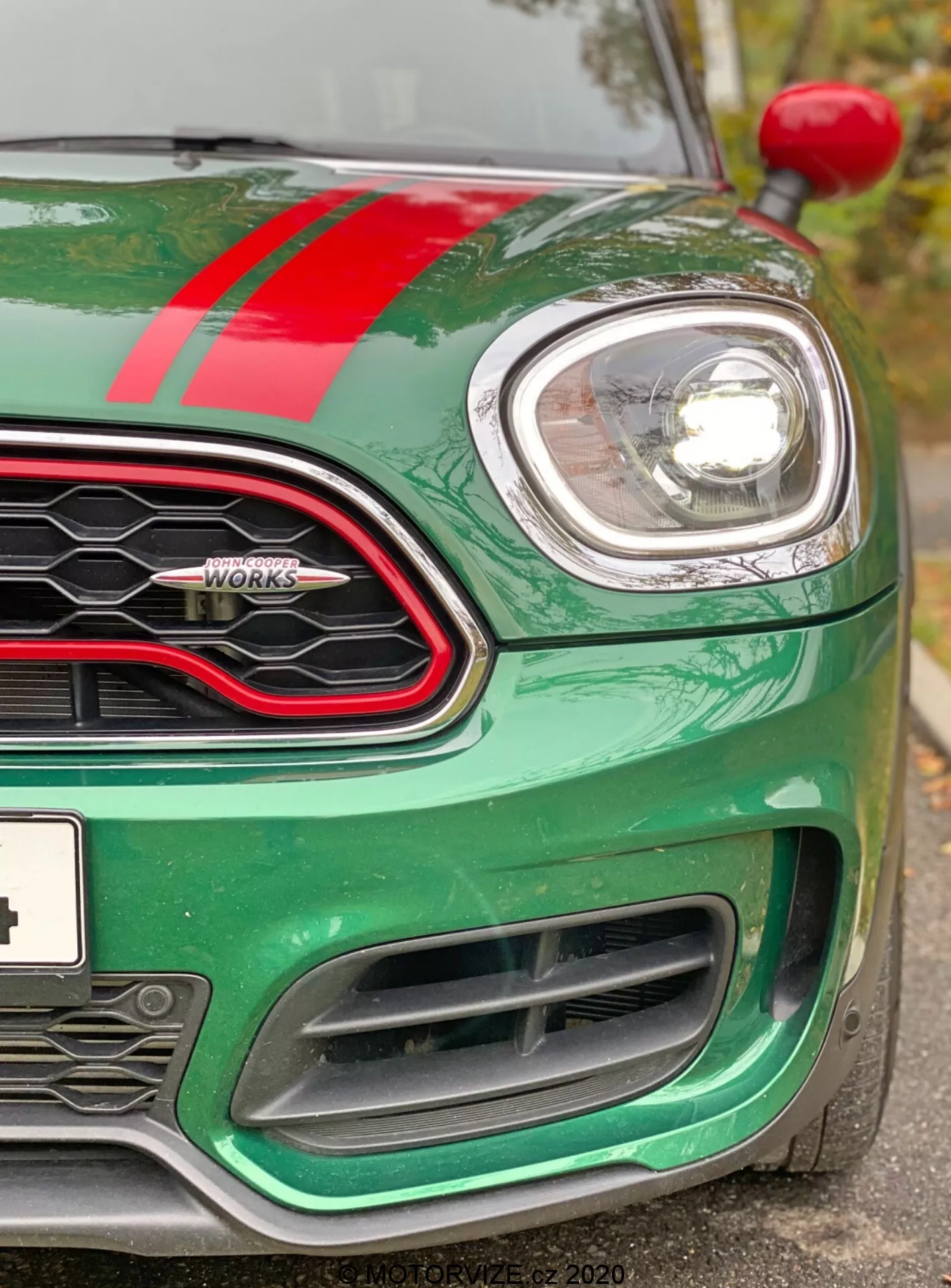 Vue en gros plan du quart avant gauche d'une Mini Countryman John Cooper Works 2020. L'image, prise légèrement en dessous du niveau des phares, met en évidence le capot vert métallisé avec une bande de course rouge, le phare gauche circulaire encadré de chrome et une partie de la grille en nid d'abeille avec une bordure noire brillante. Sont visibles l'insigne 'John Cooper Works', le clignotant gauche sur l'arche de la roue et les grandes prises d'air du pare-chocs avant conçues pour le refroidissement du moteur et la performance aérodynamique. La voiture apparaît propre et bien entretenue, soulignant sa technologie moderne de phare à faisceau projecteur ou LED et son design sportif et axé sur la performance.