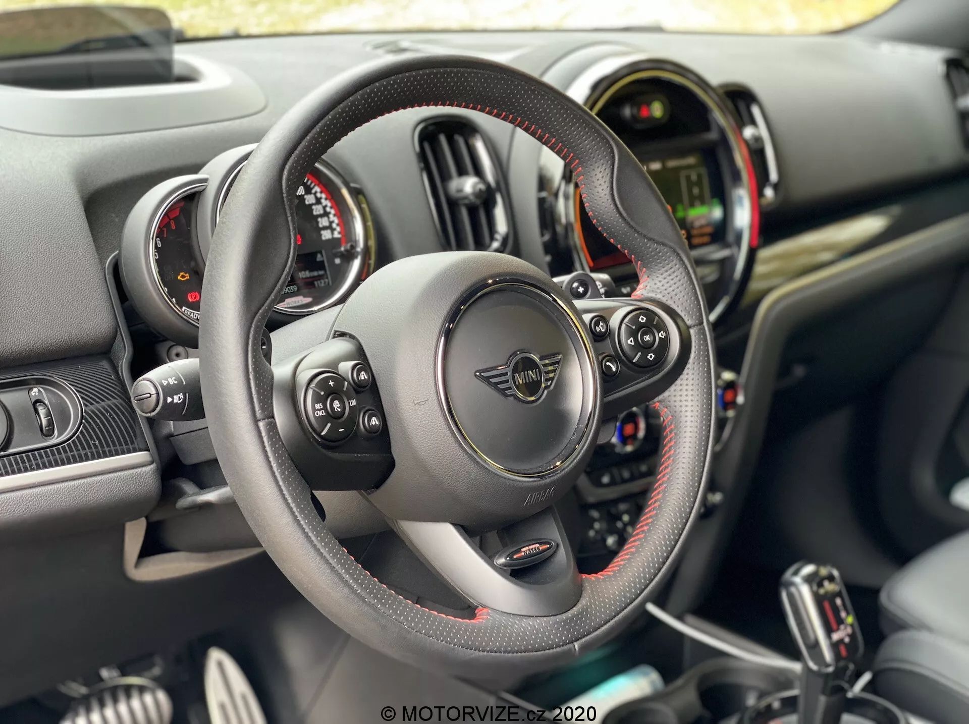 Vue intérieure de la Mini Countryman John Cooper Works 2020 depuis la perspective du conducteur, avec un volant à fond plat en cuir avec coutures visibles et commandes multifonctions, palettes de changement de vitesse, double cadrans circulaires d'instruments flanquant un écran numérique central, et un tableau de bord avec divers boutons et commutateurs sur la console centrale.
