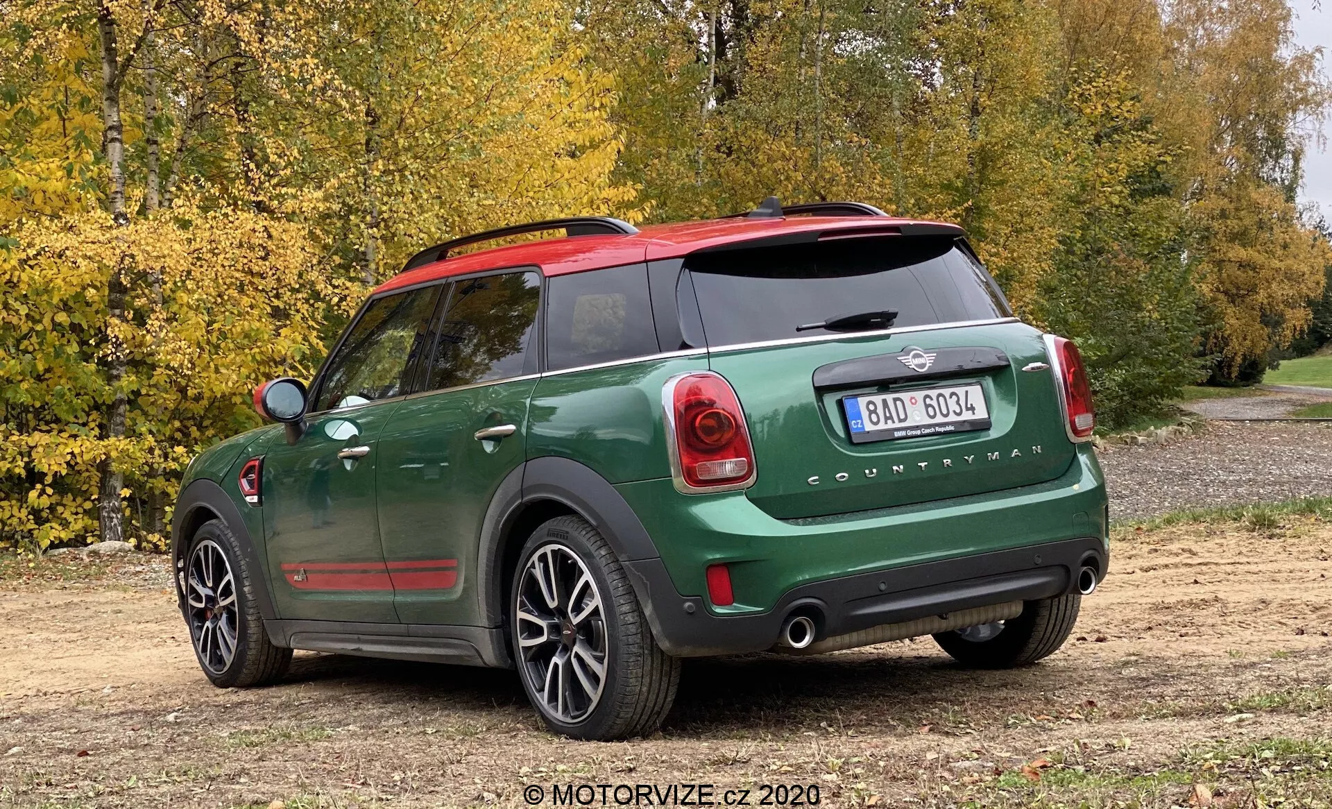 Vue arrière trois quarts d'une Mini Cooper Countryman John Cooper Works 2020 avec une peinture bicolore vert foncé et noir, des capuchons de rétroviseur rouges, un emblème Mini visible et l'inscription 'Countryman', des feux arrière au design unique, un pare-chocs arrière proéminent avec des capteurs de stationnement, une lunette arrière équipée d'un dégivreur, des doubles tuyaux d'échappement, un essuie-glace arrière, des roues en alliage à cinq branches, garée sur un chemin de terre avec des arbres et des feuilles d'automne en arrière-plan, capturée sous un léger angle élevé.
