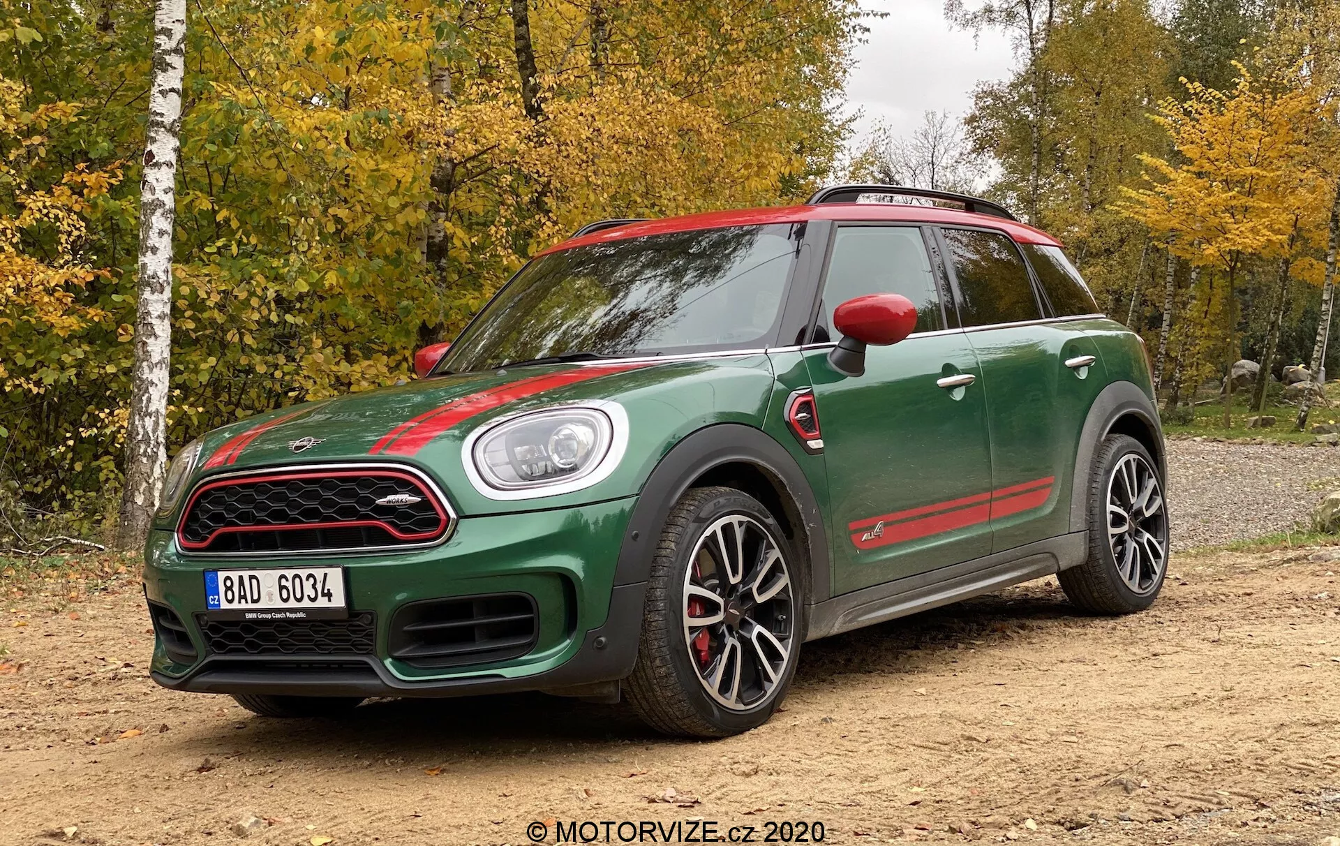 Vue de trois quarts avant d'une Mini Countryman John Cooper Works 2020, présentant une conception sportive avec une carrosserie bicolore vert foncé et des accents rouges sur les rétroviseurs, la calandre avant et les jupes latérales. Les caractéristiques comprennent une prise d'air distinctive sur le capot, des phares ronds, une calandre en nid d'abeille, de grandes roues en alliage sport avec un design multi-brins, des étriers de frein rouges, des jupes latérales prononcées et une allure agressive. La voiture est positionnée sur une surface non pavée avec une forêt en arrière-plan, sous une lumière naturelle par une journée nuageuse.