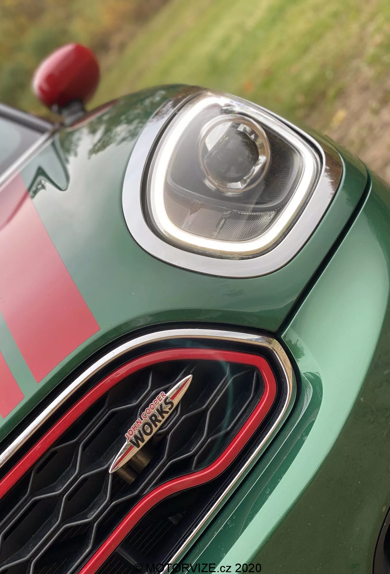 Vue rapprochée de la section avant de la Mini Countryman John Cooper Works 2020, mettant en avant le phare LED avancé et la calandre hexagonale unique avec un contour rouge et le badge 'John Cooper Works'. La carrosserie de la voiture présente une peinture verte riche avec un motif de bandes de course. Photo par PRODRIVE.CZ, 2020.