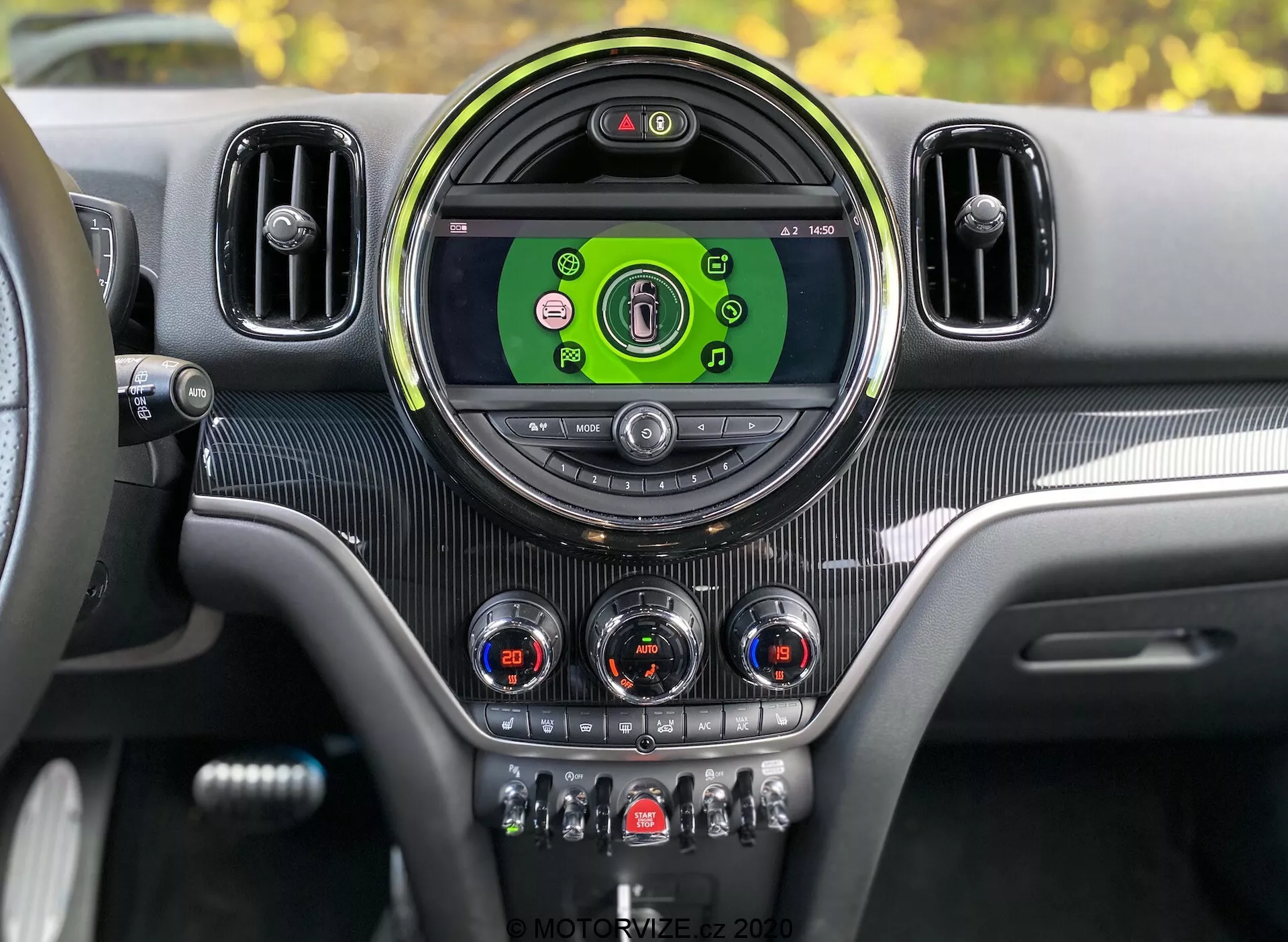 Tableau de bord intérieur du Mini Countryman John Cooper Works 2020 vu du siège conducteur. Console centrale avec grand écran circulaire d'infodivertissement affichant des icônes colorées pour les médias, la connectivité téléphonique, la navigation et les paramètres du véhicule. Encadré par une lunette circulaire élégante et flanqué de bouches d'aération supplémentaires. Sous l'écran, des cadrans analogiques avec accents chromés pour le contrôle du climat et des interrupteurs pour des fonctions comme le chauffage des sièges, le dégivrage, la recirculation de l'air et les modes de conduite. Texture de type fibre de carbone et accents métalliques offrent un design intérieur moderne et luxueux avec un tableau de bord numérique visible indiquant que le véhicule est allumé.