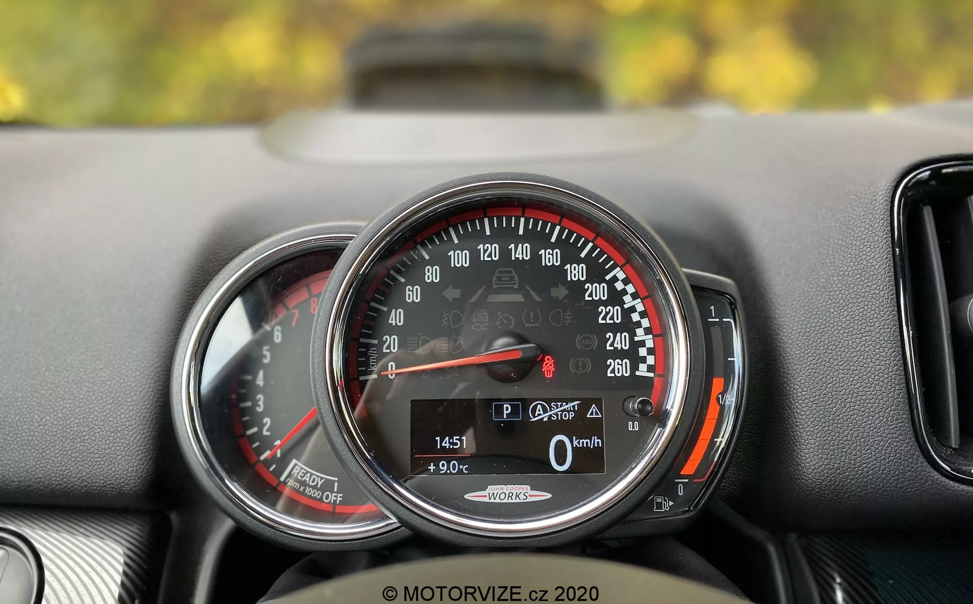 Gros plan détaillé sur le tableau de bord du Mini Countryman John Cooper Works 2020. Le focus est mis sur un compteur de vitesse circulaire allant de 0 à 260 km/h, avec l'aiguille indiquant 0 km/h, ce qui signifie que la voiture est à l'arrêt. Un affichage numérique en bas montre la voiture en mode 'P', une température extérieure de 9,0°C, un kilométrage de 1451 km et une icône de coffre ouvert. Les mots 'Auto Start/Stop' suggèrent un système d'économie de carburant. Le compteur de vitesse est encadré par un anneau chromé et a une zone rouge à partir de 220 km/h. À gauche, une vue partielle d'un autre indicateur, probablement un compte-tours. Le tableau de bord a une surface texturée et une petite bouche d'aération est partiellement visible à droite du compteur de vitesse.