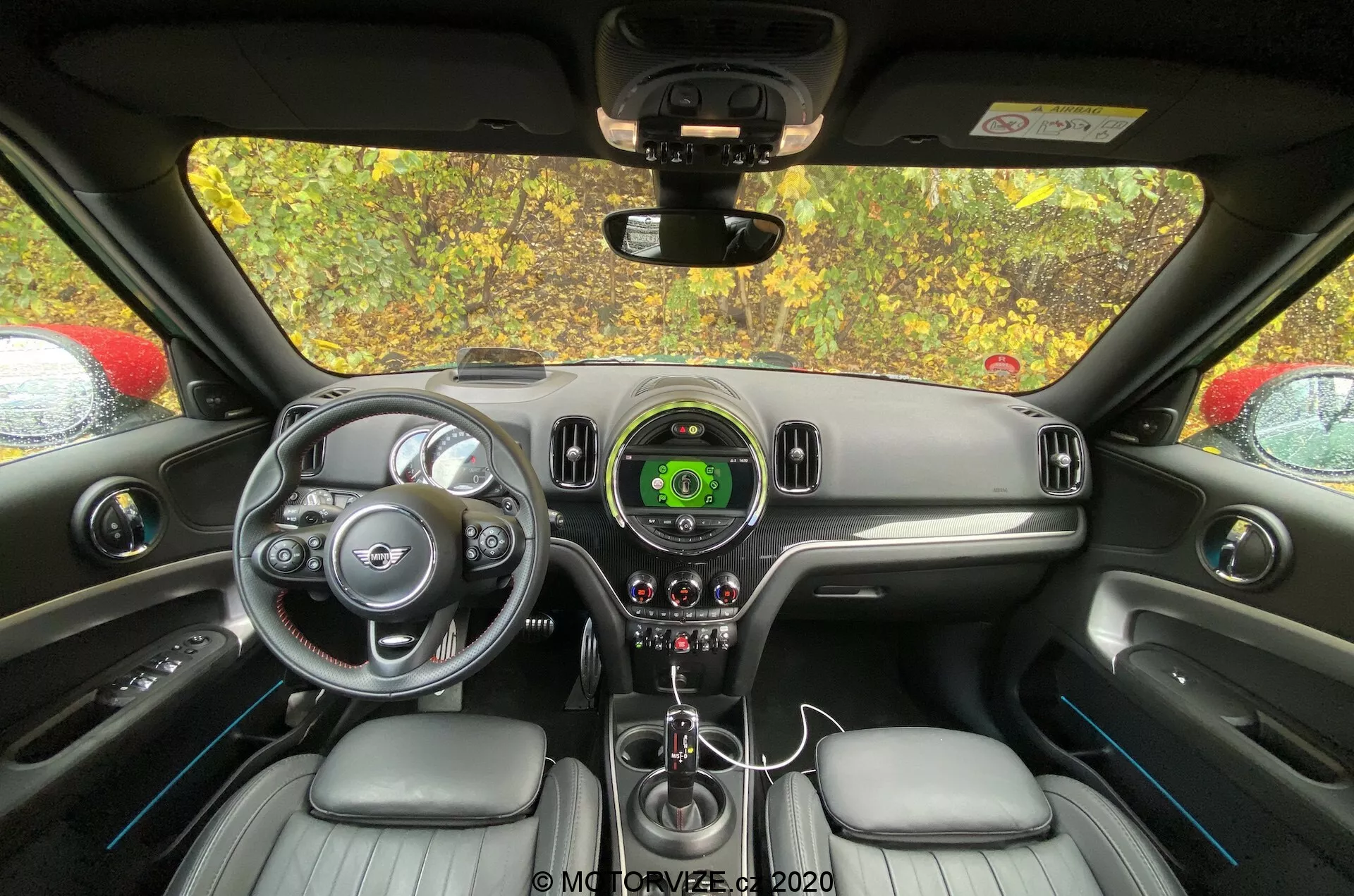 Intérieur du Mini Countryman 2020 John Cooper Works vu depuis la perspective du siège arrière, avec un focus sur le tableau de bord et la zone de commande du conducteur. Le volant avec accents argentés et logo de la marque, le compteur de vitesse central avec affichage numérique, le design élégant du tableau de bord, le levier de vitesses, les contrôles du climat, les sièges revêtus de cuir et le rétroviseur sont visibles.